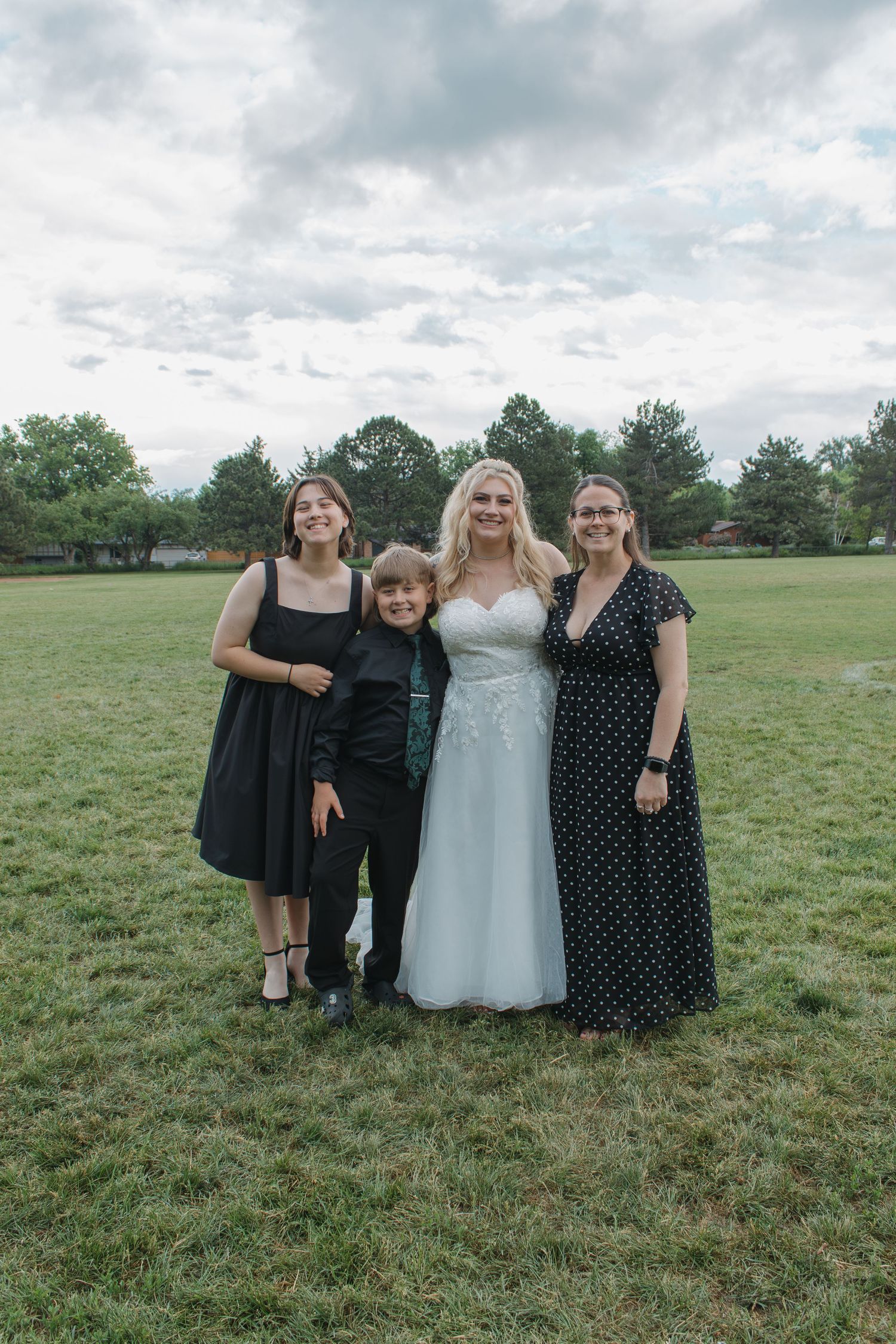 Small wedding group photo on grass field with bride in white gown and guests in dark dresses.