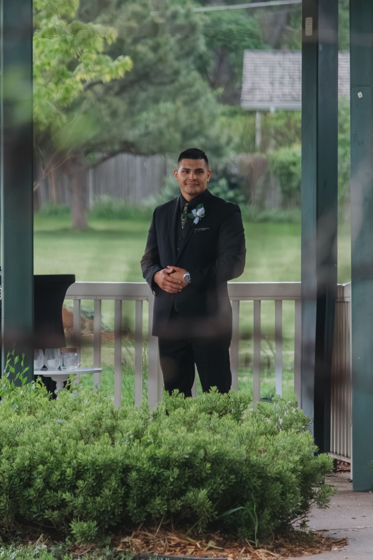 A person in formal attire stands on a gazebo deck surrounded by landscaped bushes.