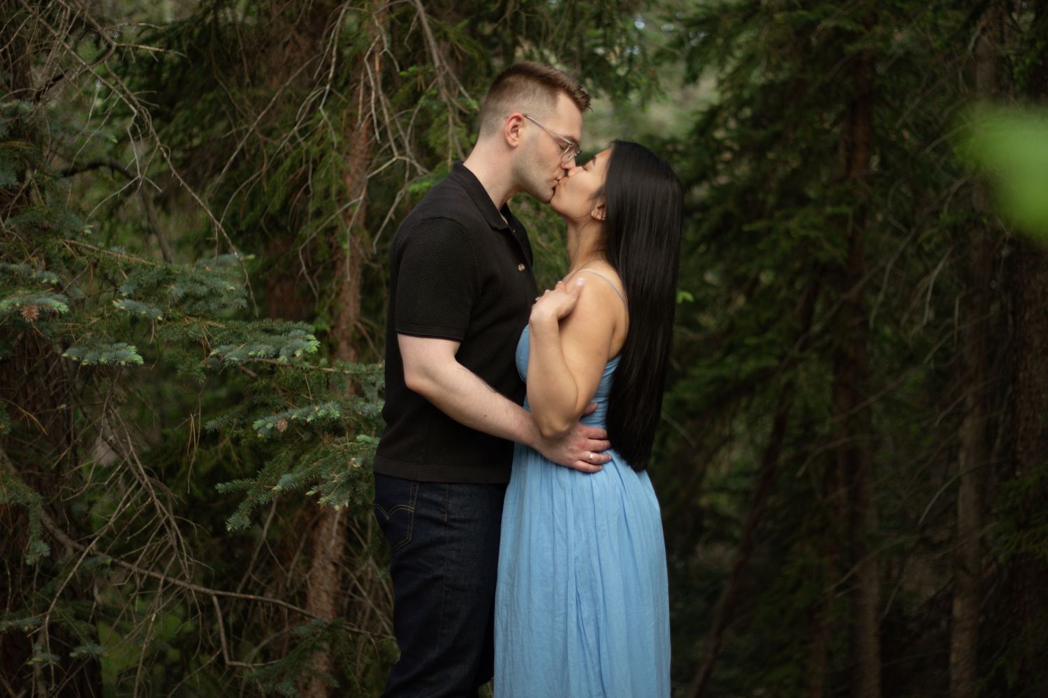 A romantic couple shares an intimate moment in a wooded setting with green foliage surrounding them.