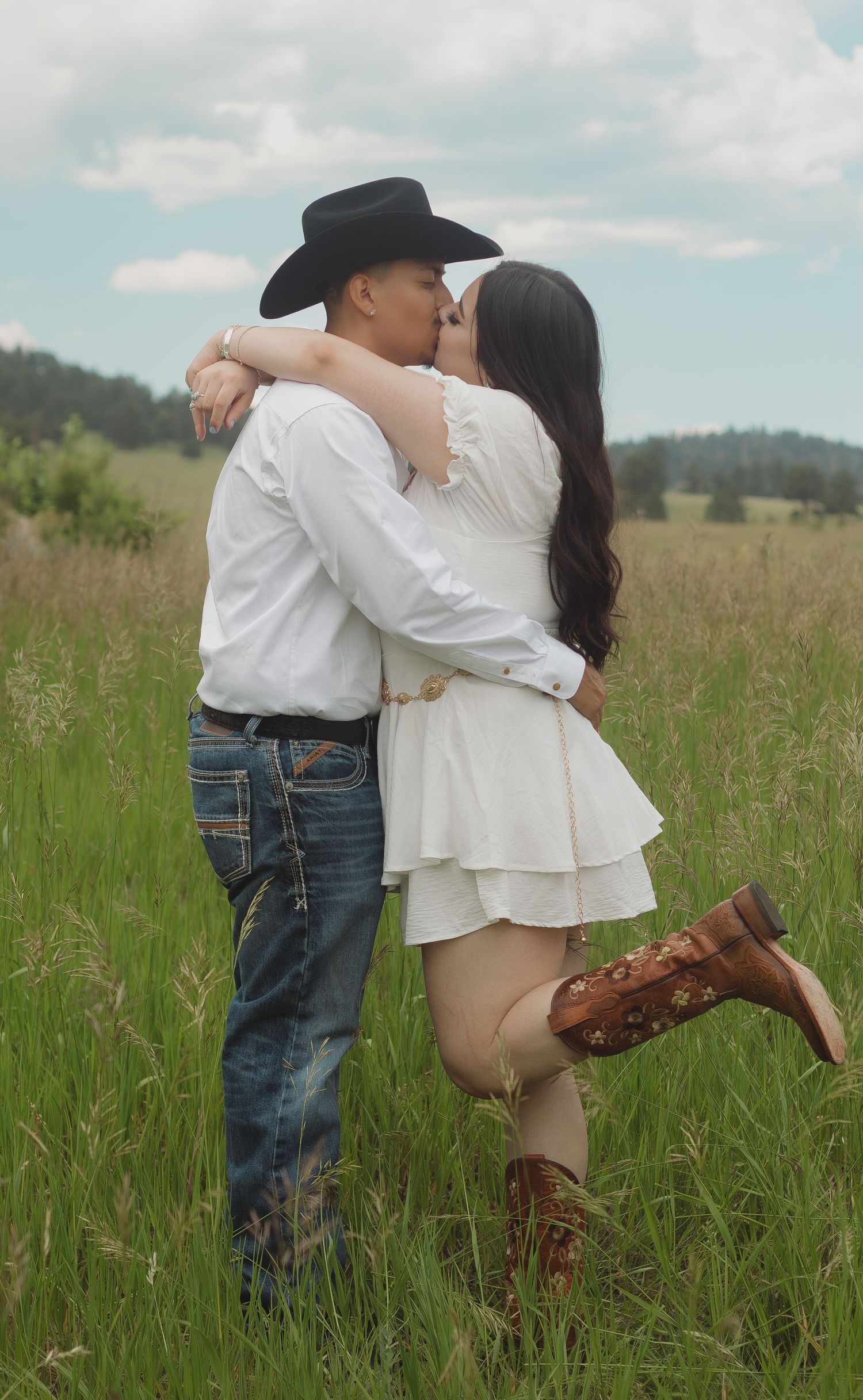 Romantic countryside embrace with cowboy hat and boots against mountain backdrop.