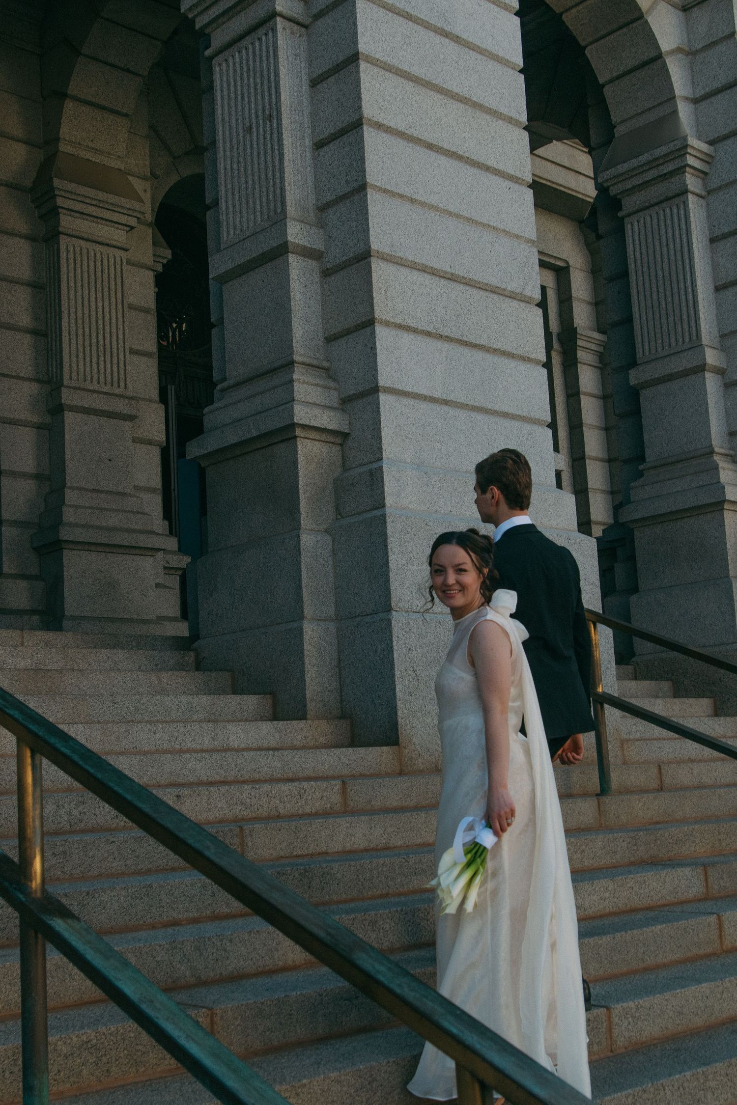 A couple in wedding attire walks up grand stone steps with classical columns in the background.