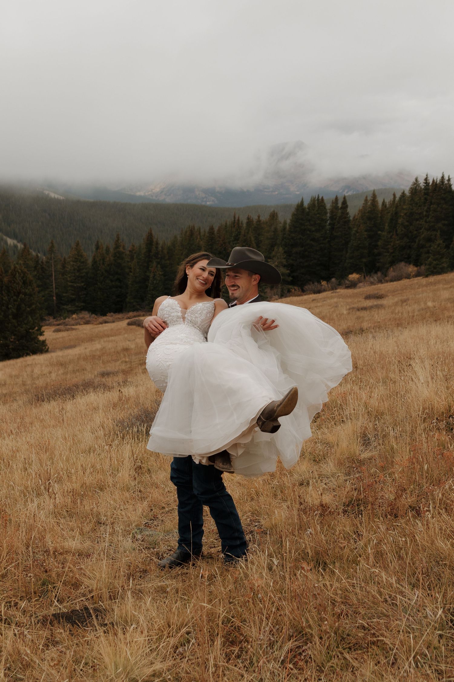 A romantic couple poses in a mountain meadow with dramatic cloudy skies and evergreen trees in the background.