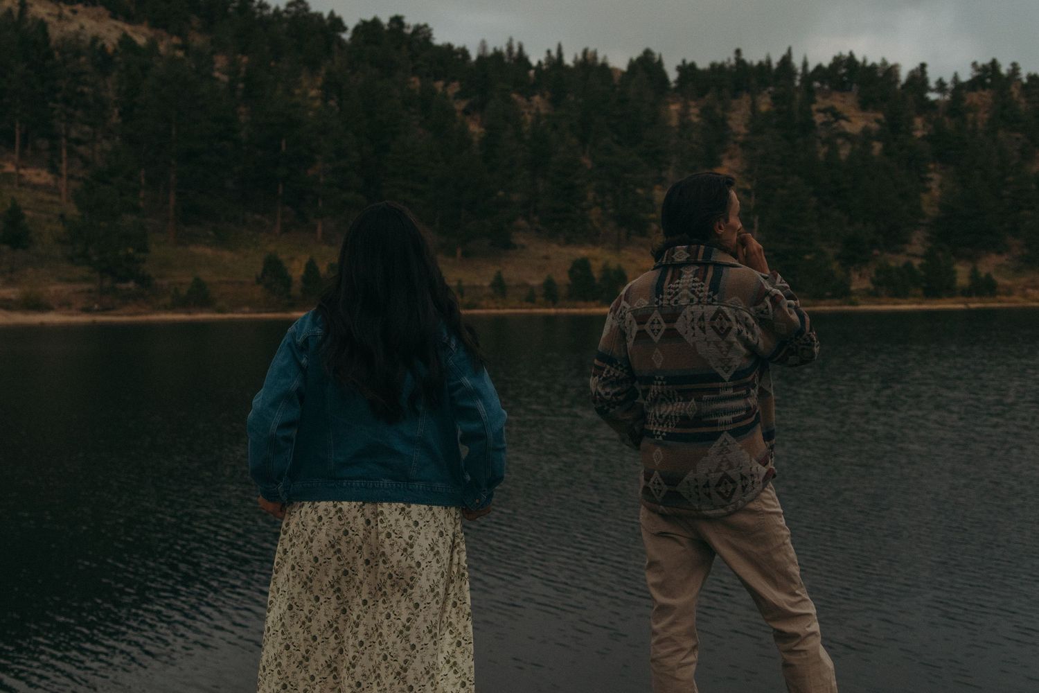 A couple stands together at the edge of a tranquil lake surrounded by evergreen trees and mountains at sunset.