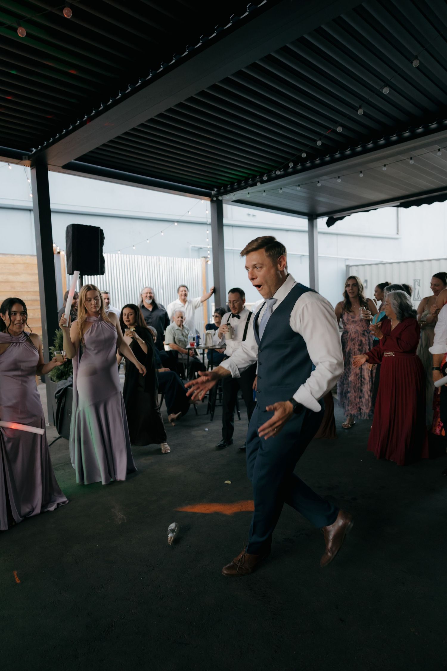 Groom in navy vest dancing energetically on dance floor surrounded by cheering wedding guests.