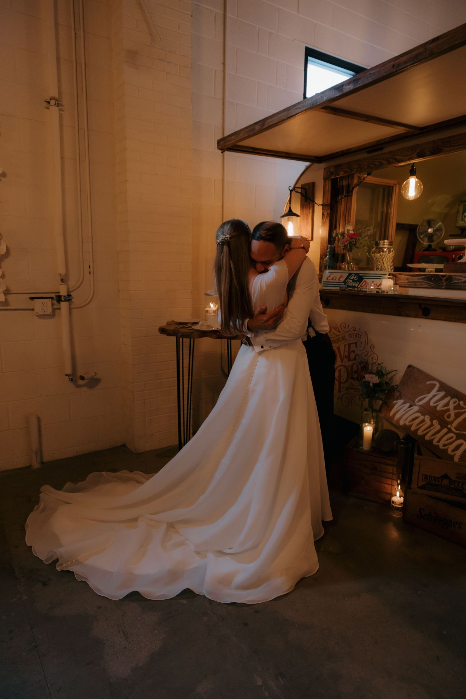 Bride and groom sharing romantic embrace near candlelit bar with Just Married sign at wedding venue.