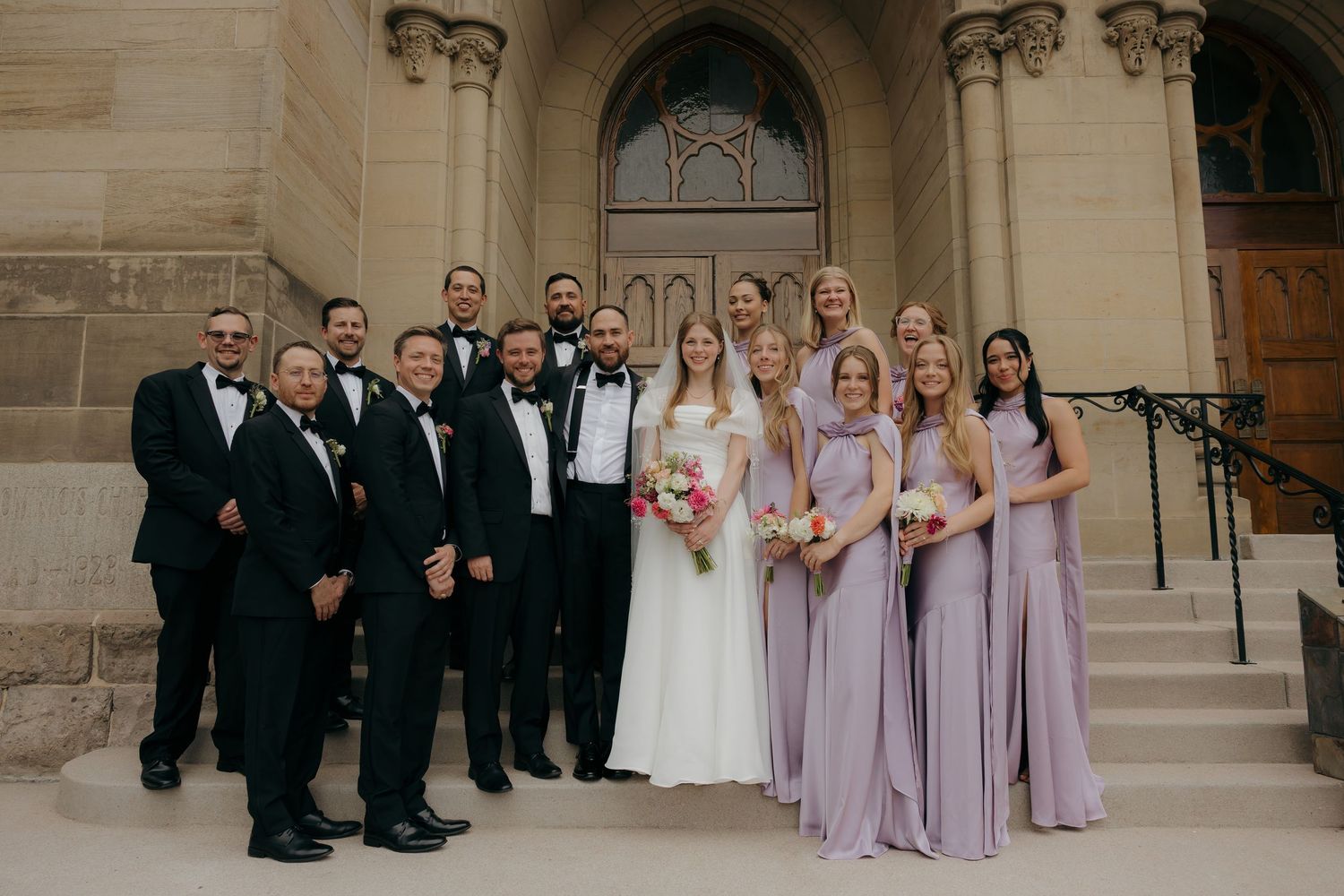 Wedding party posing on church steps, bridesmaids in lavender gowns and groomsmen in black tuxedos.