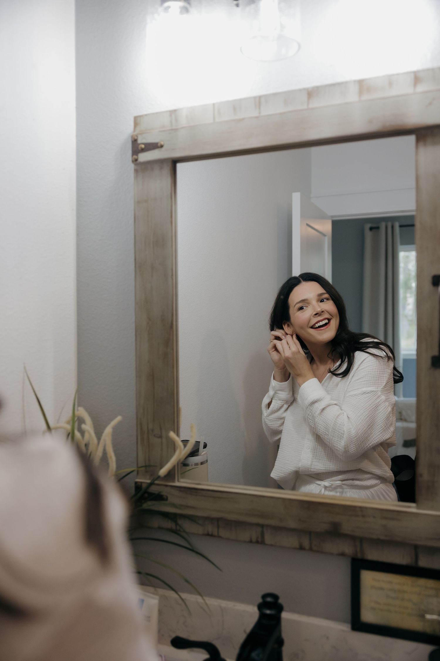 A smiling woman in a white robe puts on earrings while looking at her reflection in a rustic wood-framed bathroom mirror.
