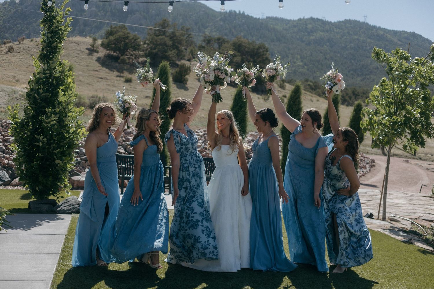 Bride and bridesmaids in blue floral dresses raising bouquets outdoors at a hillside wedding venue.