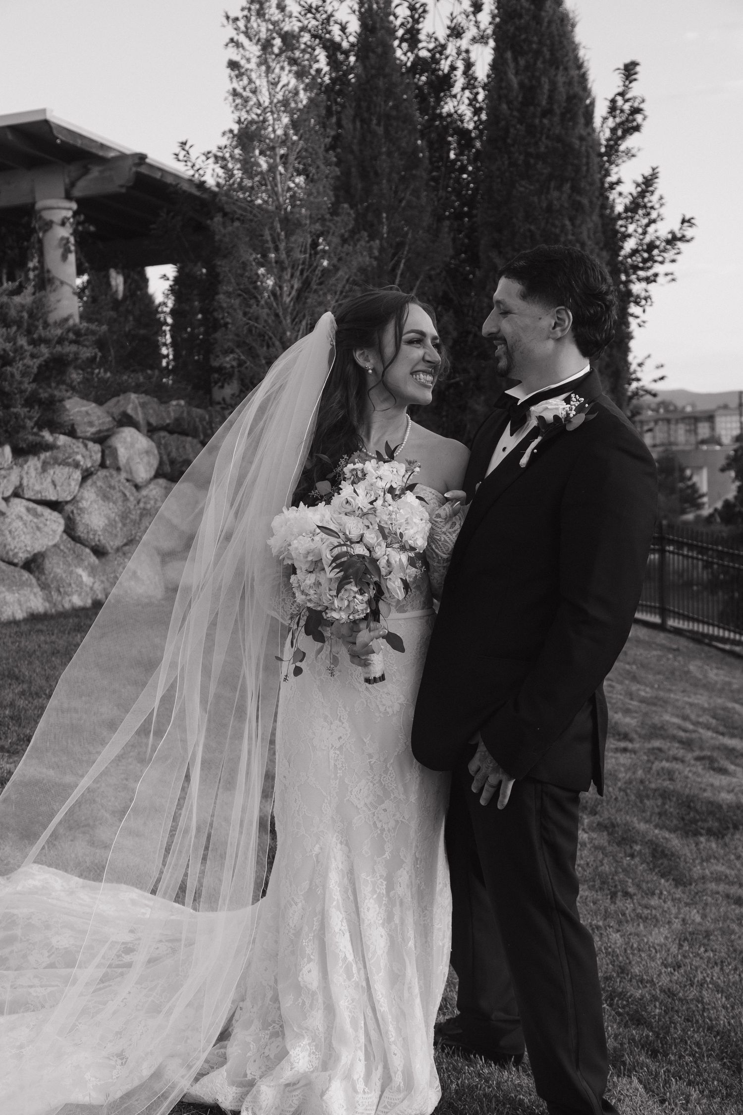 Black and white photo of smiling bride and groom in wedding attire standing on grass with veil flowing.