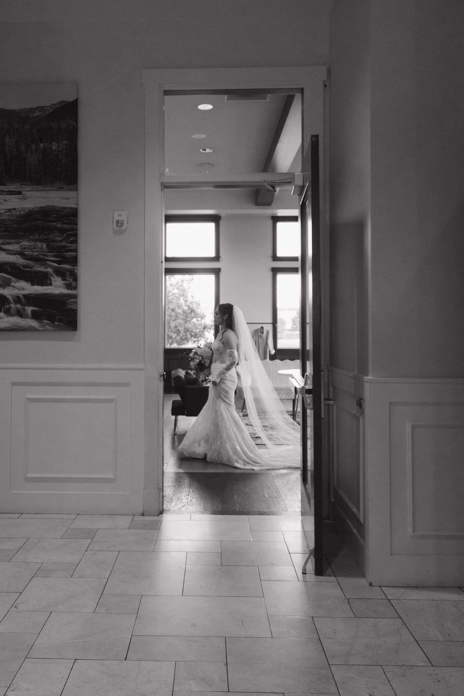 Elegant bride in wedding gown and veil holding bouquet framed in doorway, black and white photo.