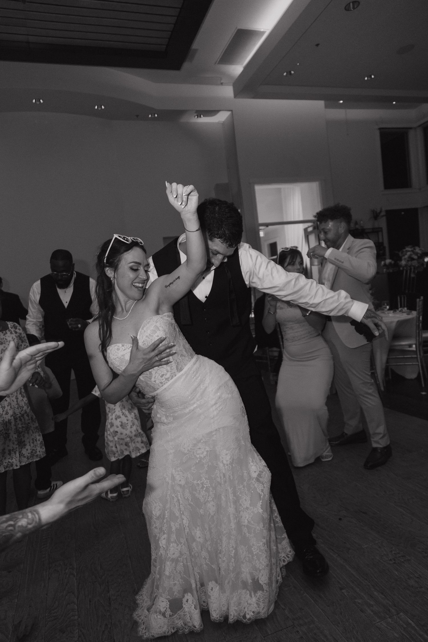 Black and white photo of bride and groom dancing energetically with guests at wedding reception.