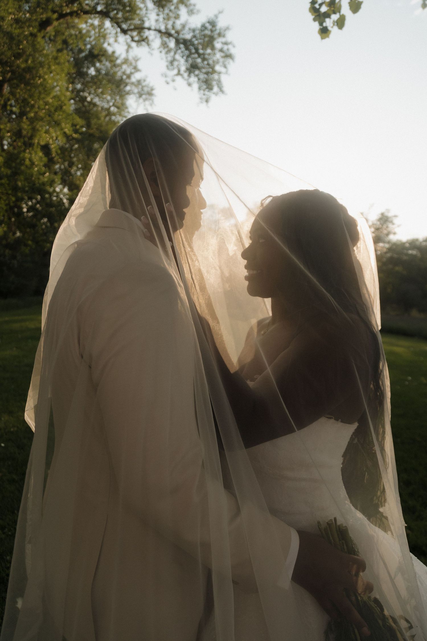 Bride and groom share an intimate moment under a sheer veil with warm backlit sunlight in an outdoor setting.