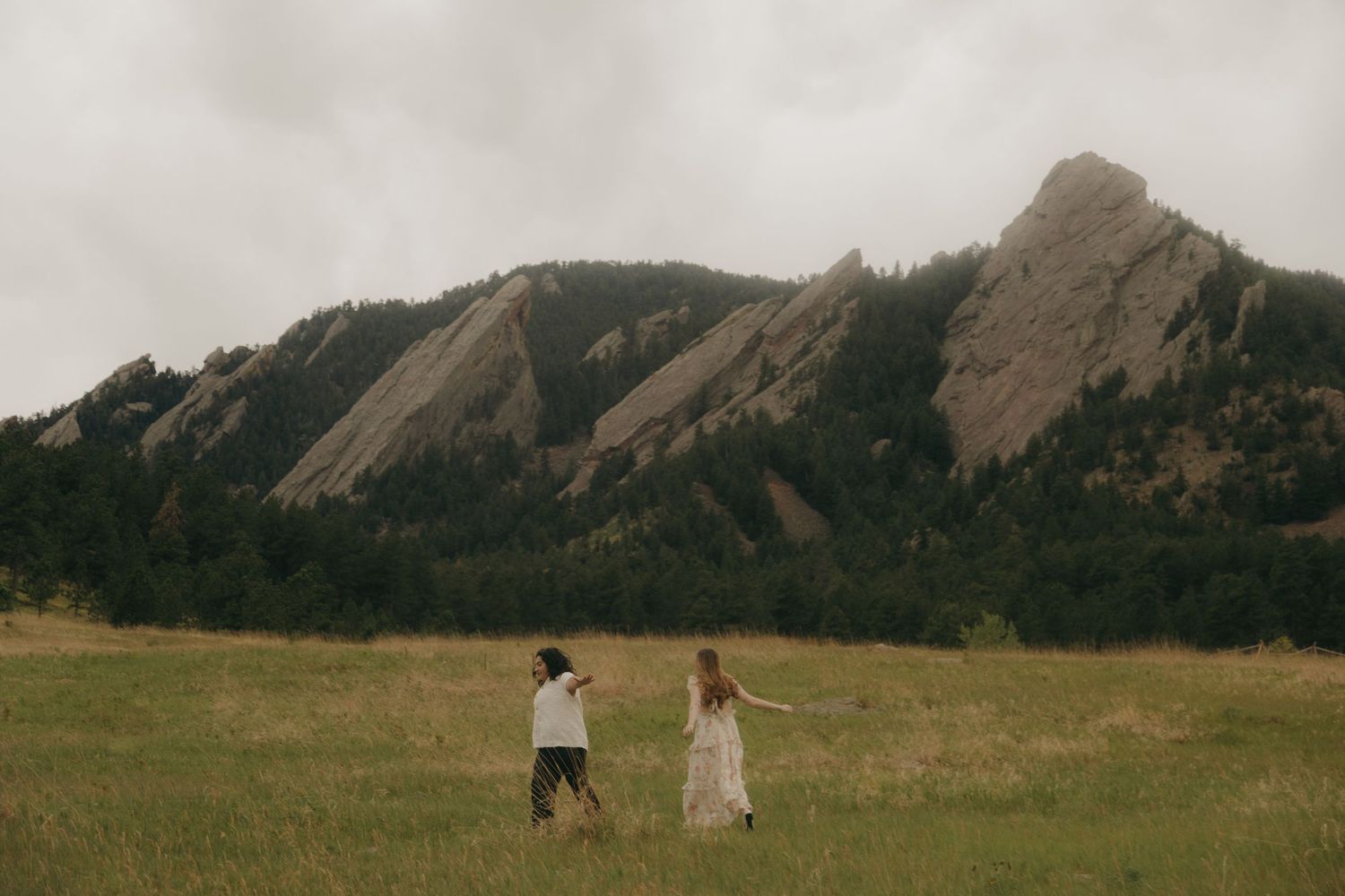 Couple walking hand in hand through a grassy meadow with dramatic Flatirons rock formations behind.