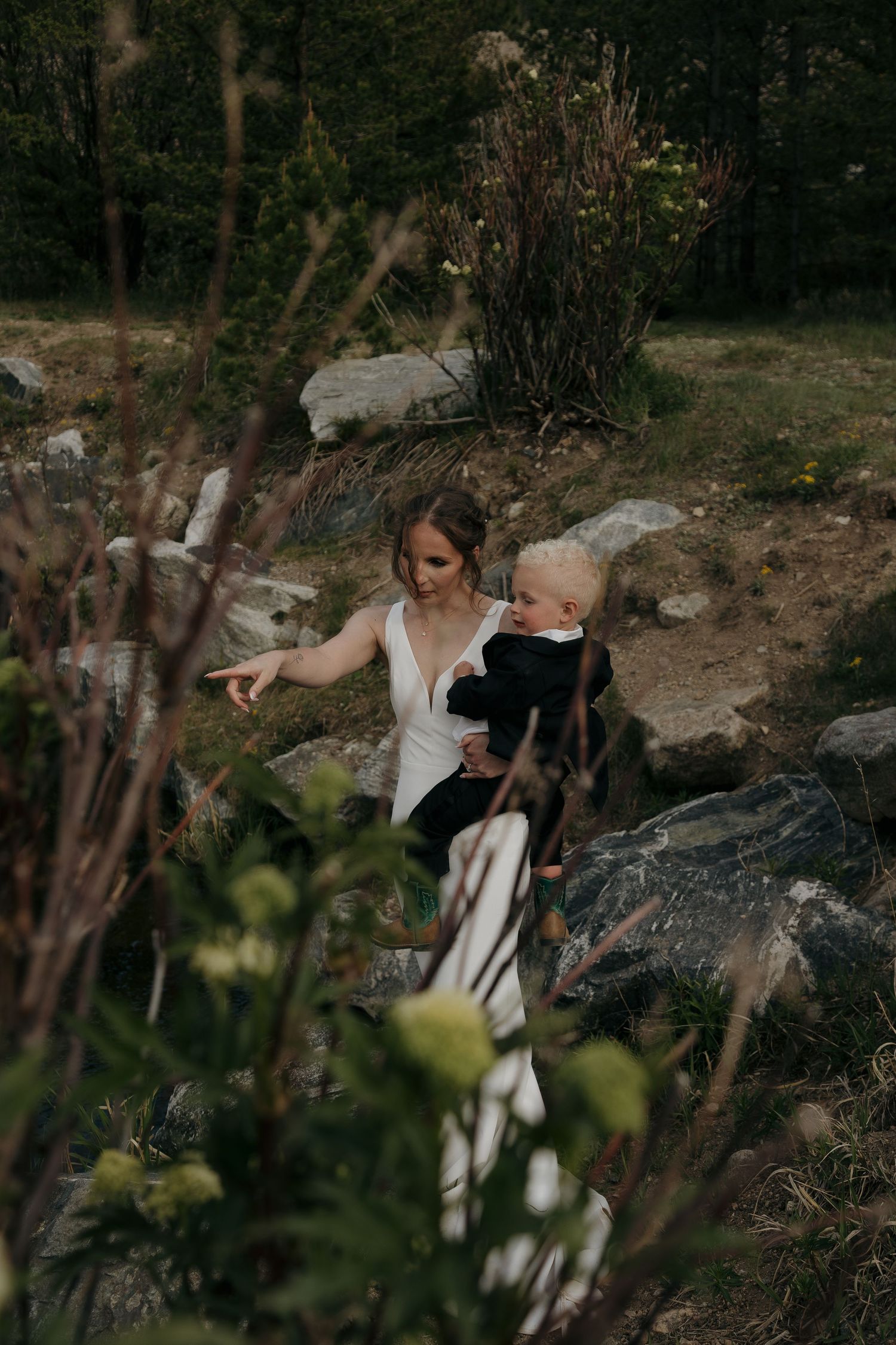 Bride in white wedding dress holds toddler while exploring rocky garden landscape.