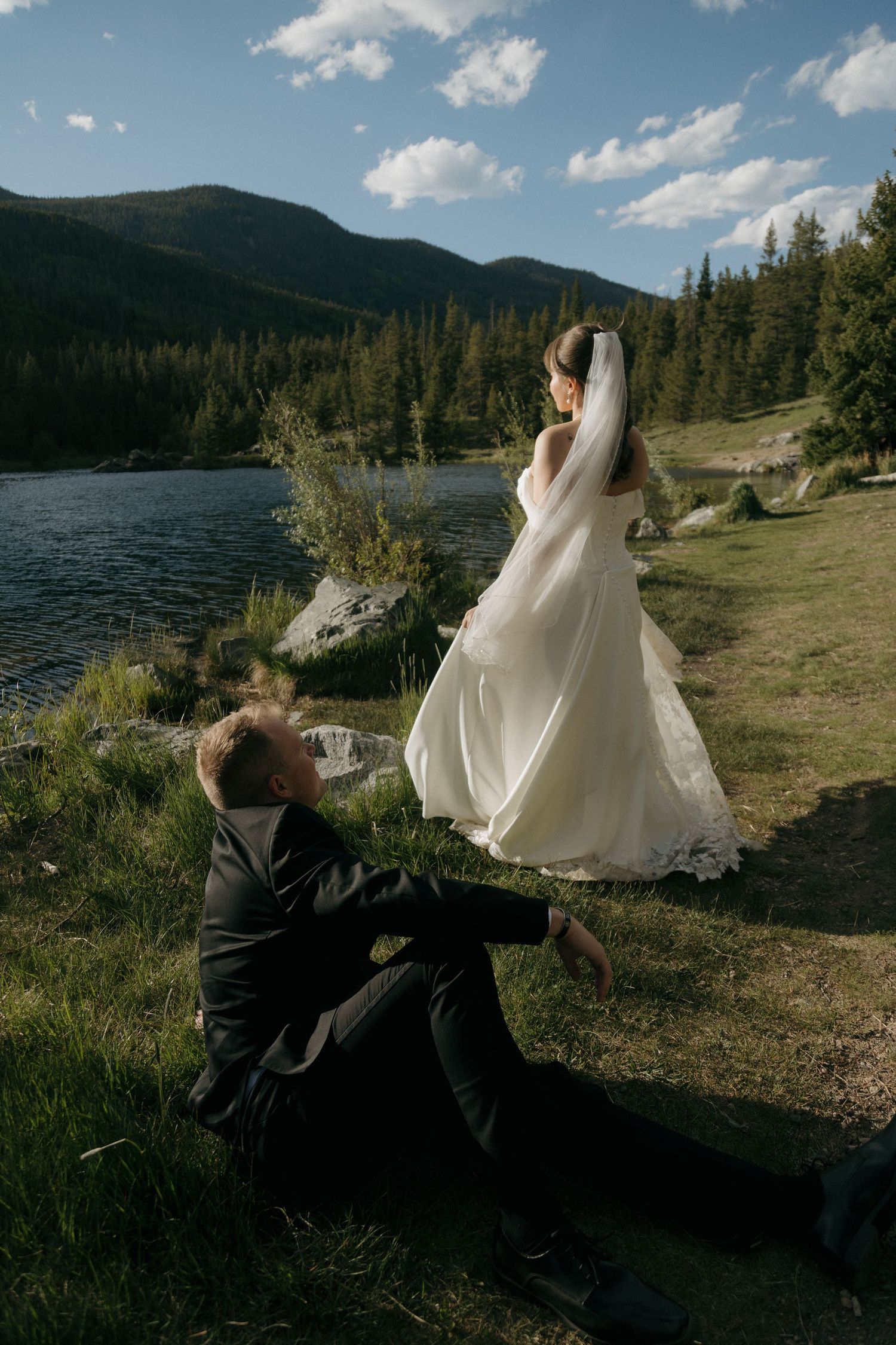 Bride in flowing white gown and veil standing by mountain lake while groom sits on grass.