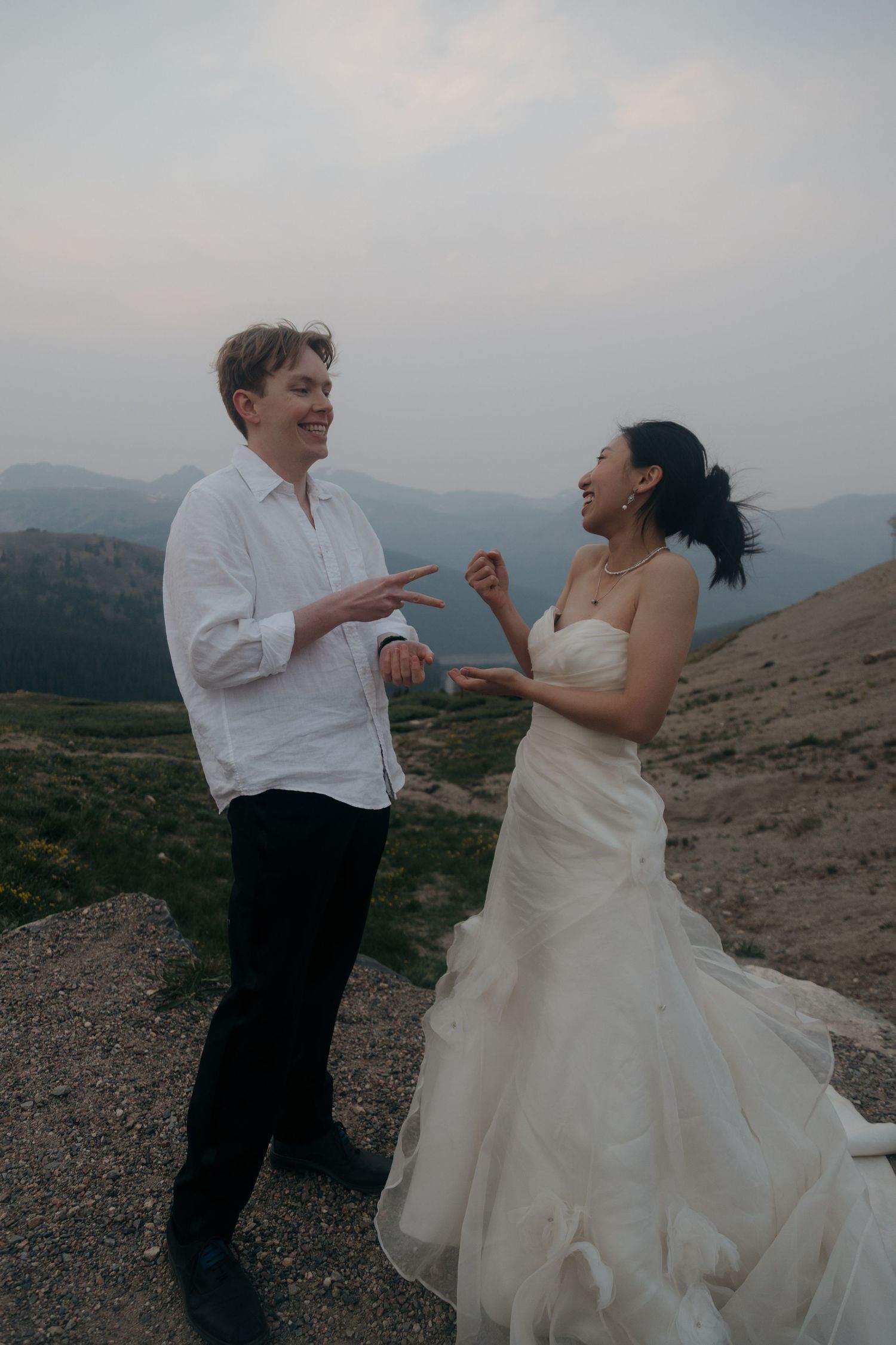 Bride and groom laugh joyfully together on a mountain ridge, her white gown flowing in the wind at sunset.
