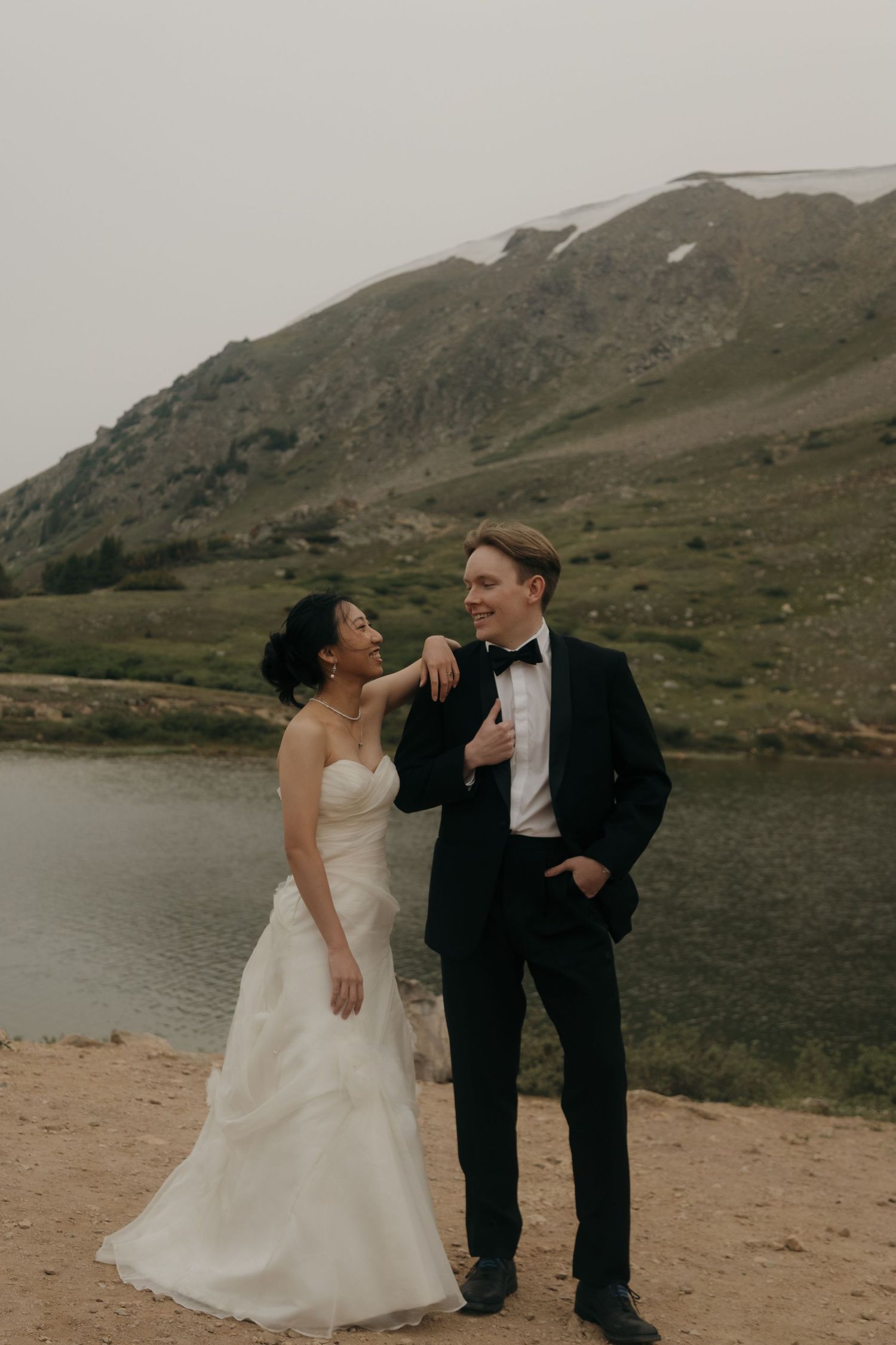 Bride and groom share a joyful moment by a mountain lake, she in a strapless gown, he in a black tuxedo.