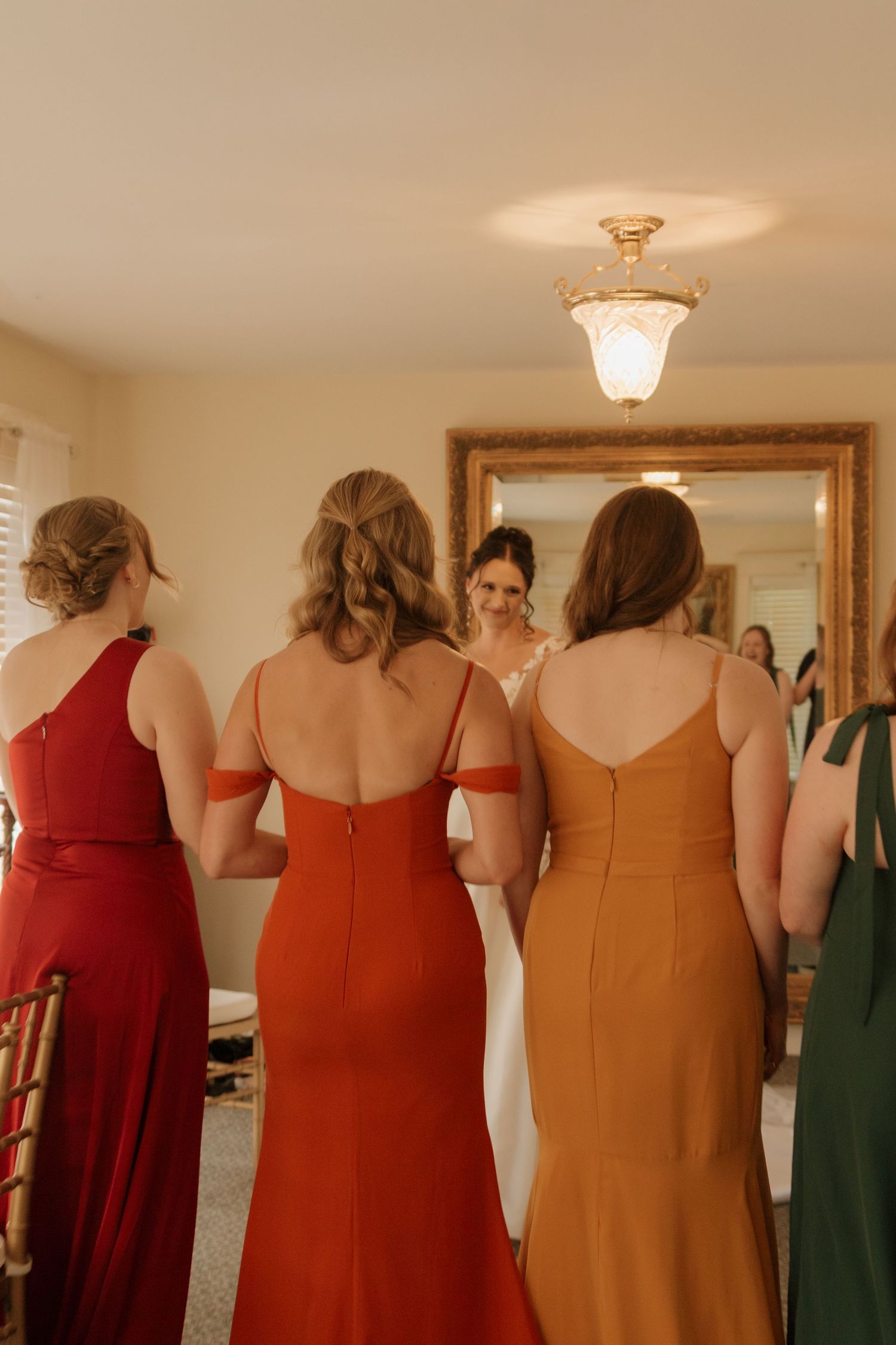 Bridesmaids in rust and burgundy dresses stand in a row showing the back details of their gowns.