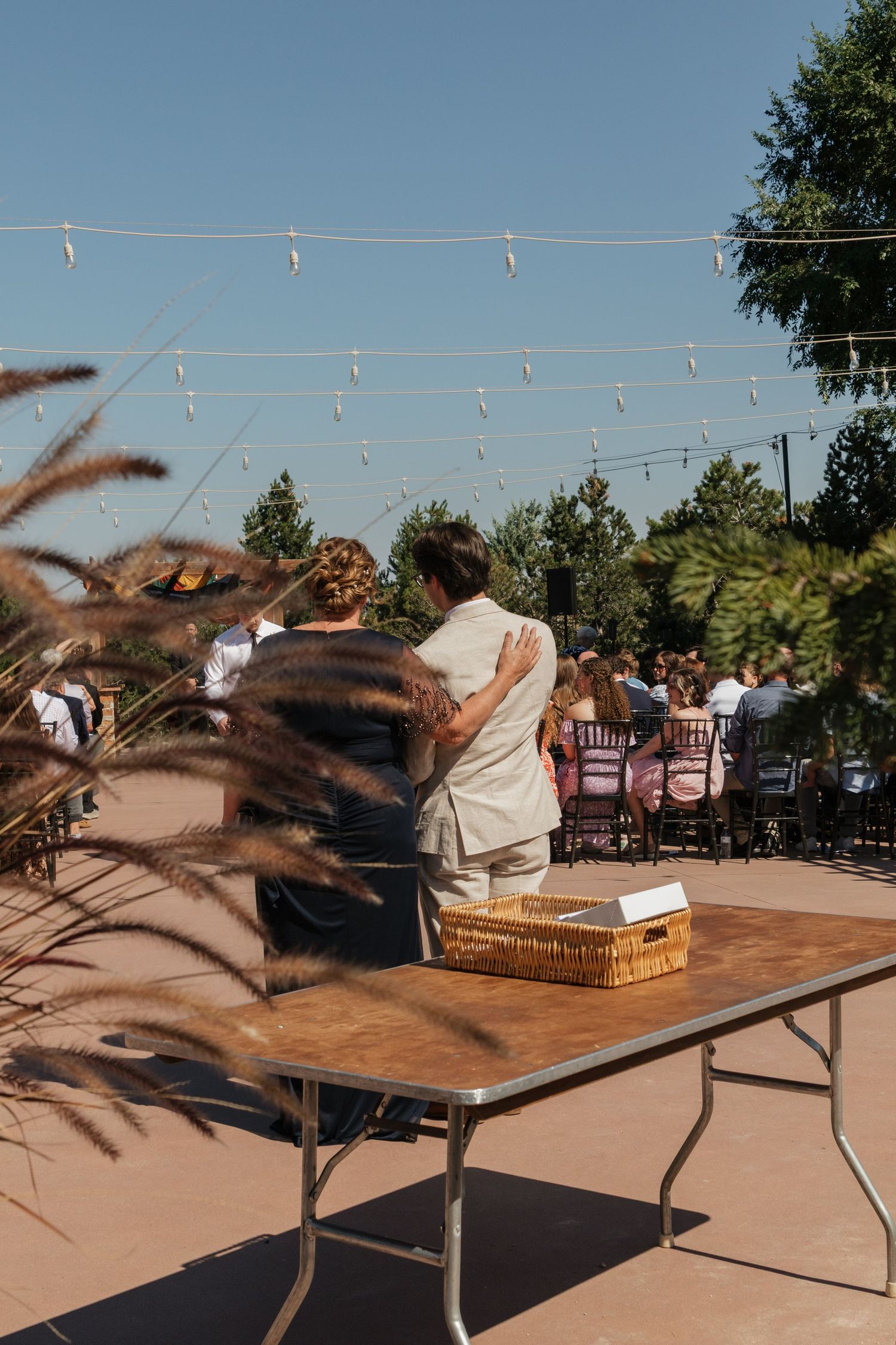 Outdoor wedding reception setup with string lights and wooden tables against a sunset sky.