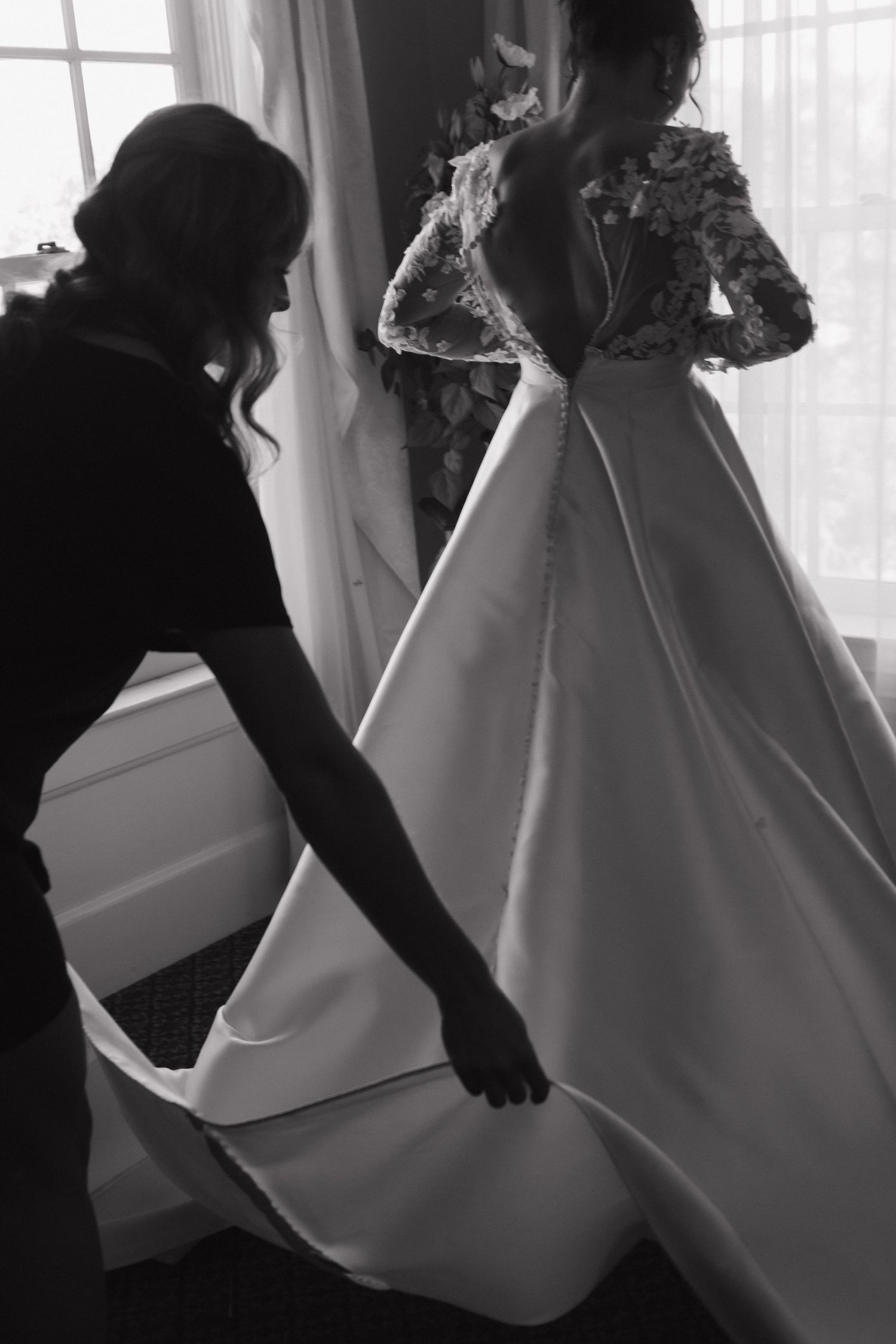 Black and white silhouettes adjust a flowing wedding dress in natural window light.