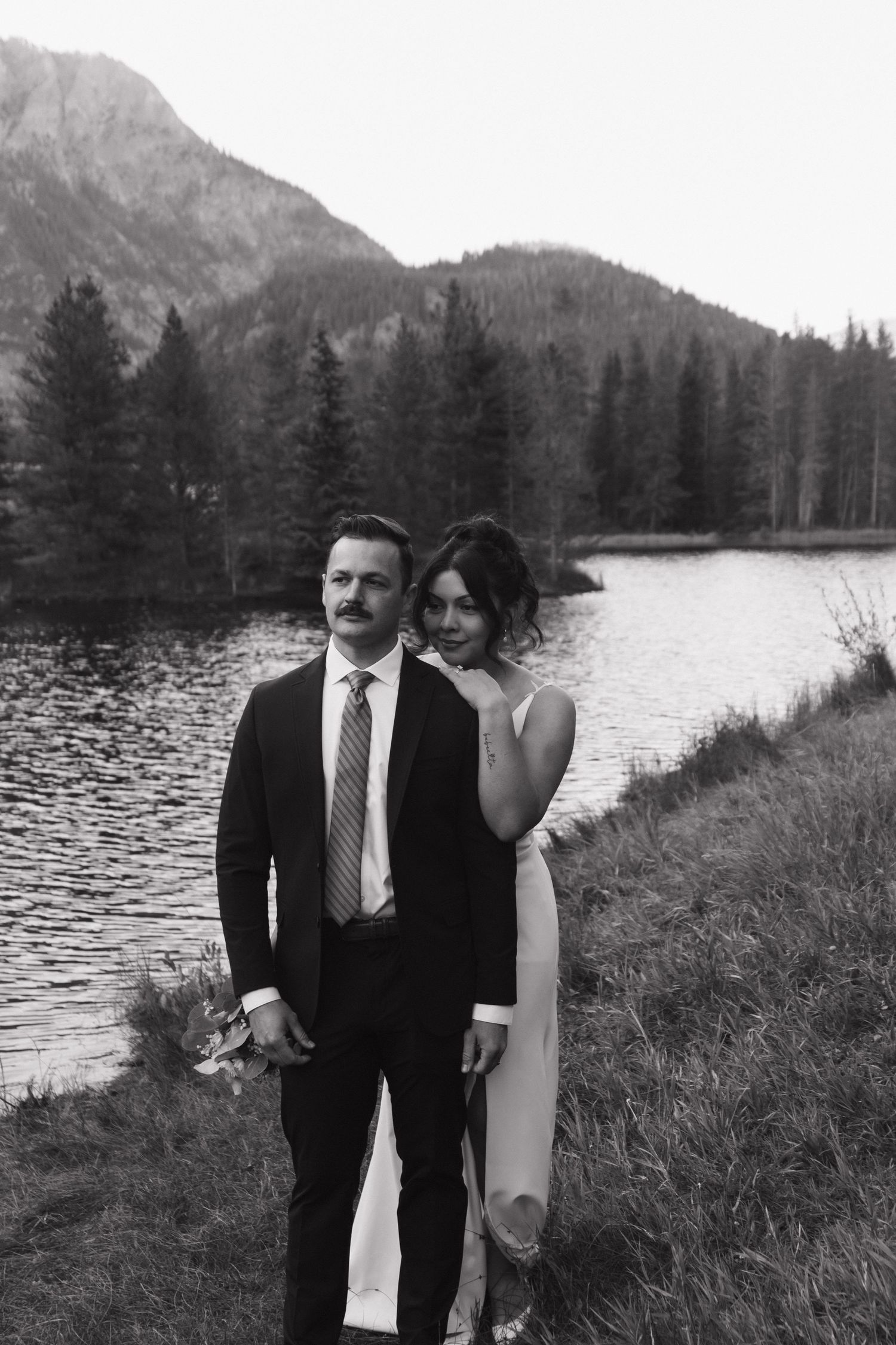 A romantic wedding couple poses by a mountain lake surrounded by evergreen trees in black and white.