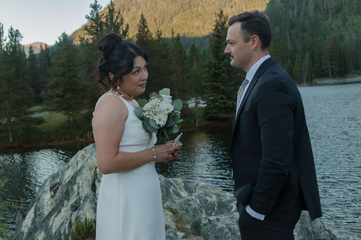 A wedding couple shares a moment by a mountain lake with a bouquet of white flowers.