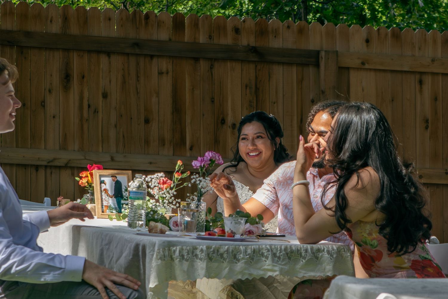People enjoying an outdoor garden party with wine glasses around a decorated table in sunlit backyard setting.