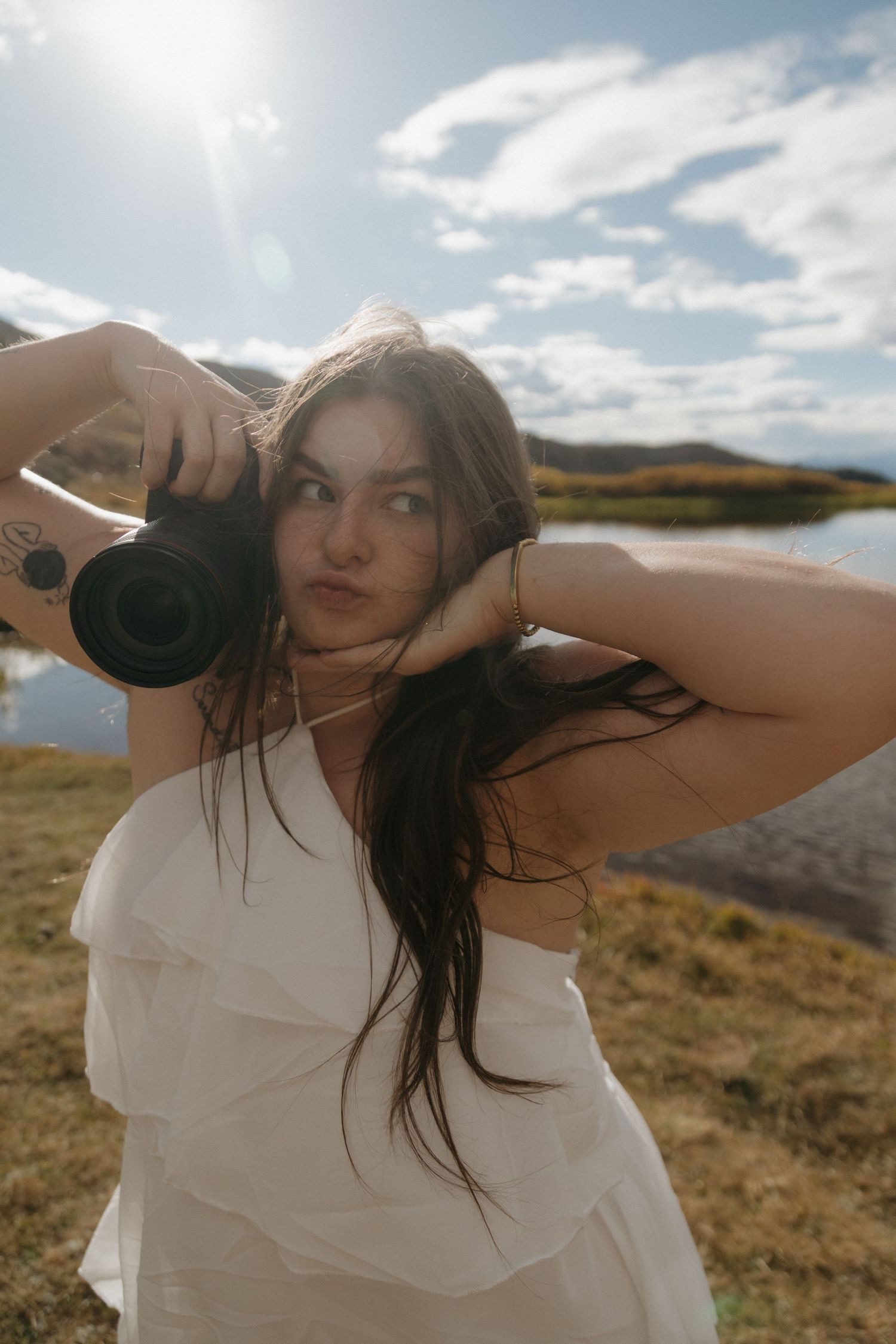 Young woman in white dress holding camera outdoors near lake, sunlit sky and hills in background.