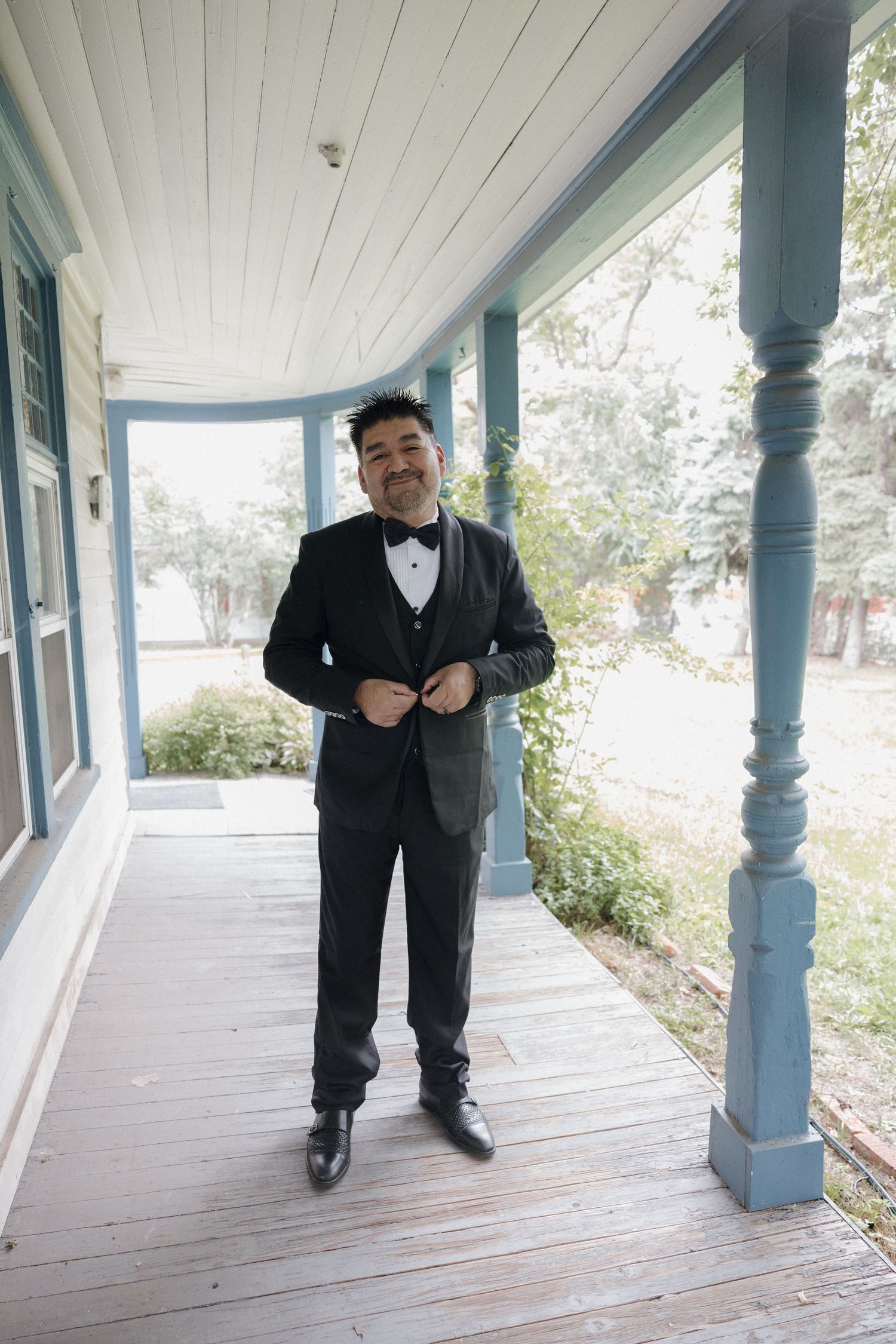 Smiling groom in black tuxedo with bow tie buttoning jacket on vintage blue porch.