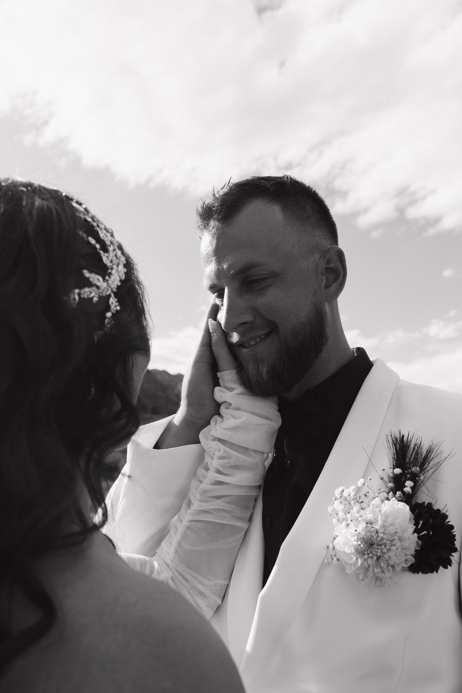 Black and white wedding photo of groom smiling as bride tenderly touches his face, both in elegant attire outdoors.