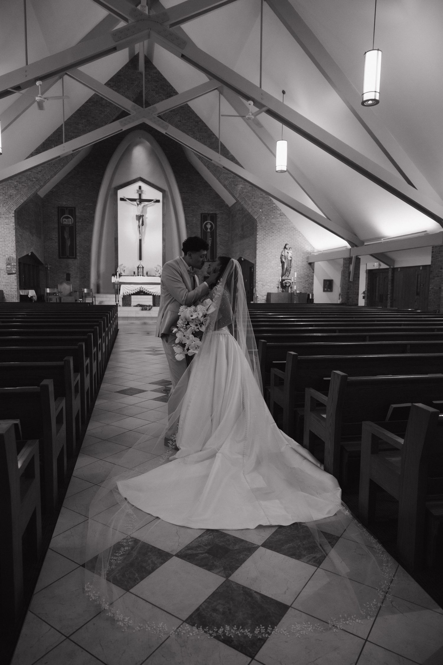 Black and white photo of a bride and groom sharing a kiss in a church aisle, with pews and a crucifix altar in the background.