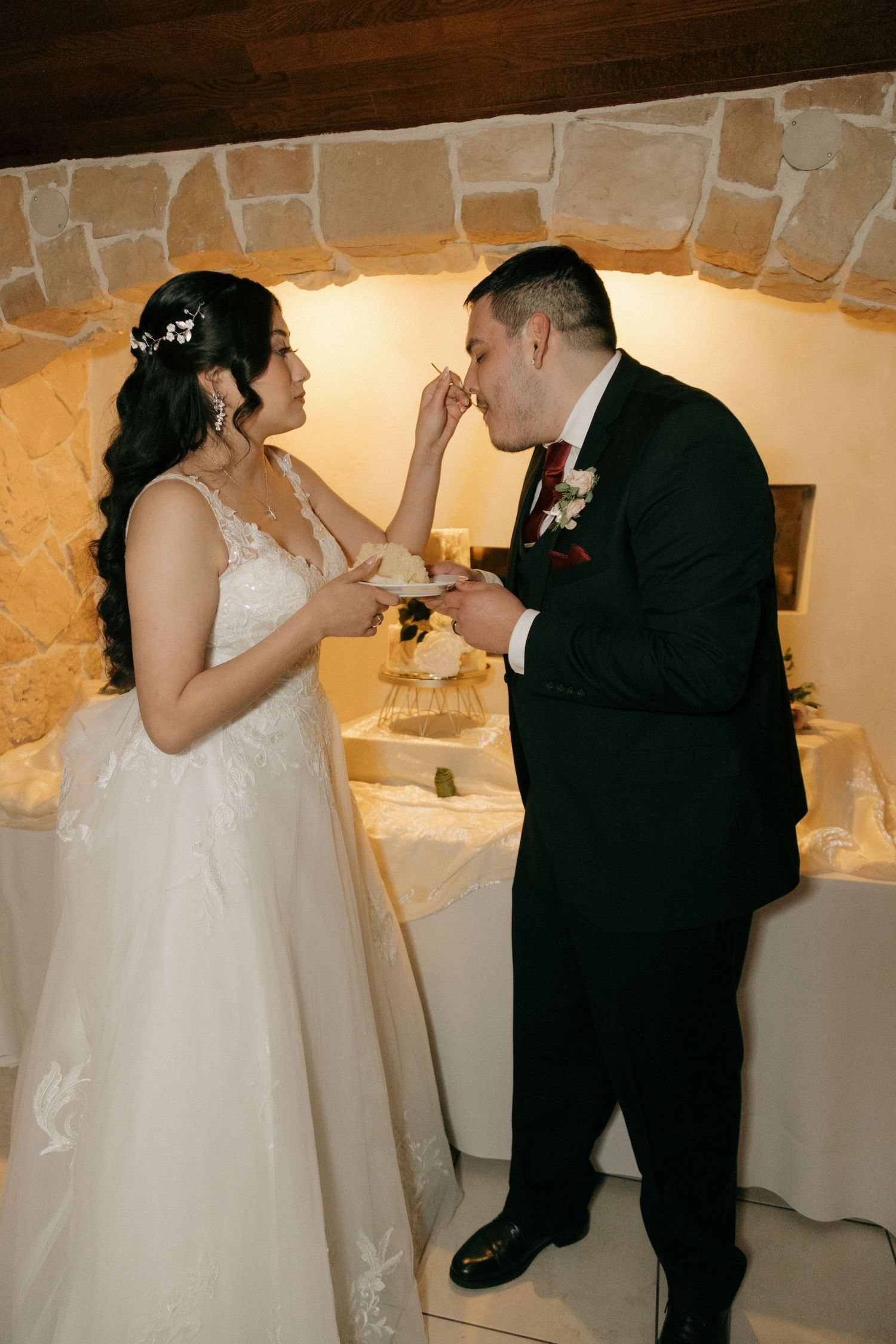 Bride feeds groom wedding cake during reception in rustic stone-walled venue with warm lighting.