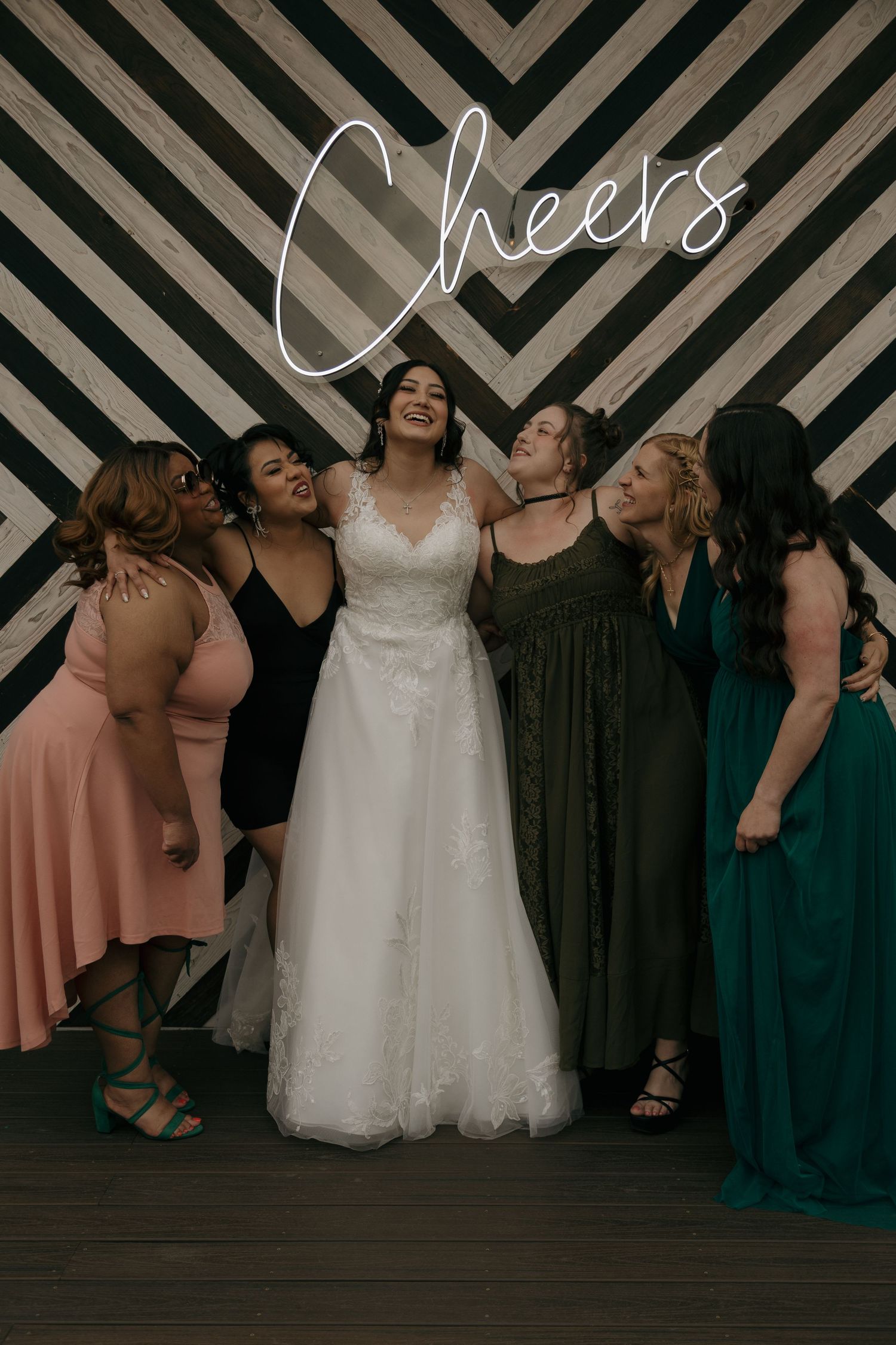 Bride in white lace gown poses with five friends under a Cheers neon sign at wedding photo booth.