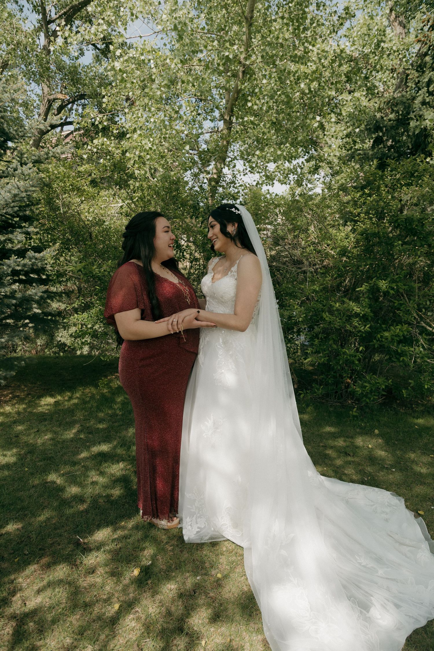 Bride in white gown and veil holds hands with bridesmaid in red dress outdoors among green trees.