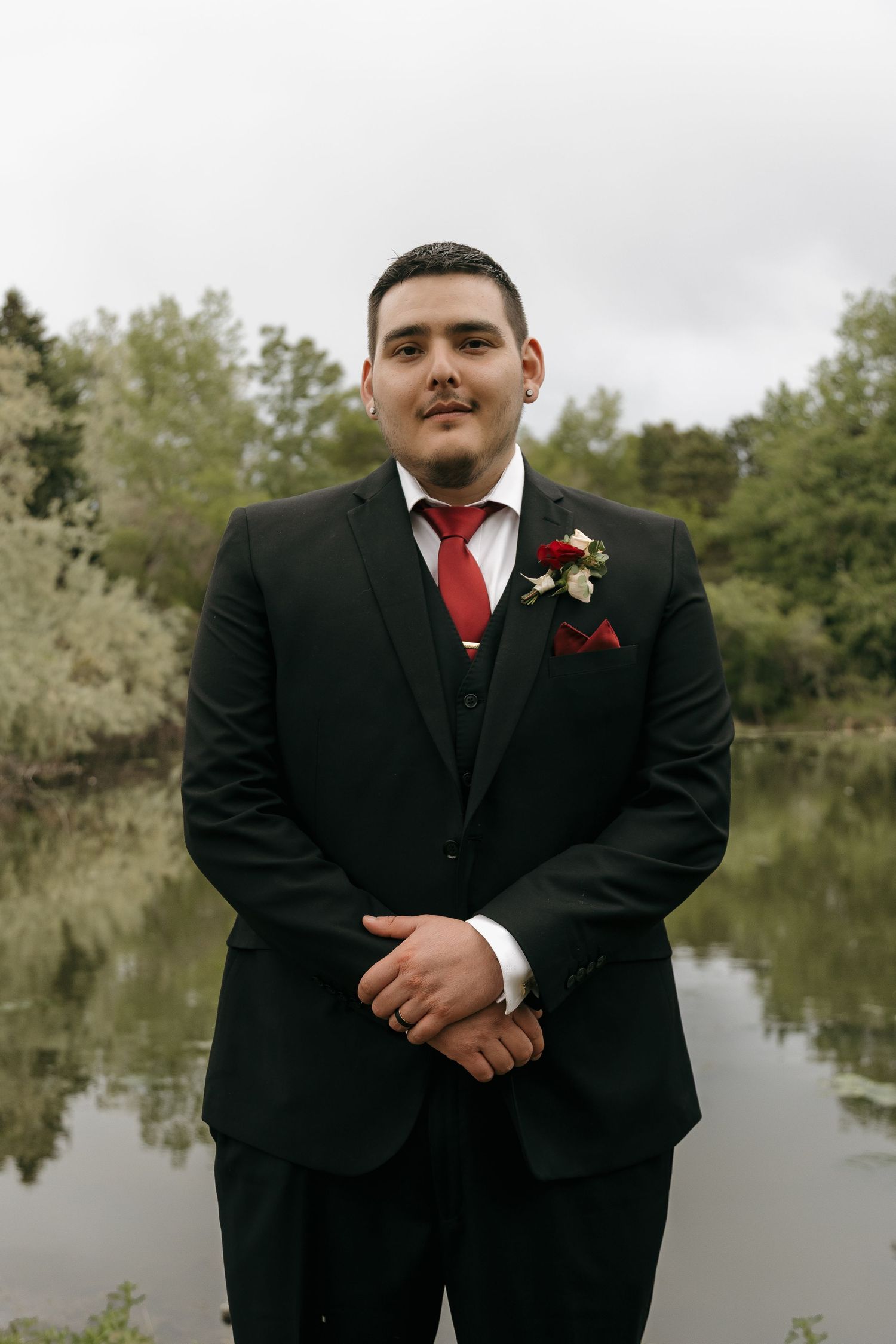 Dapper groom in black suit with red tie and floral boutonniere poses by a serene pond.