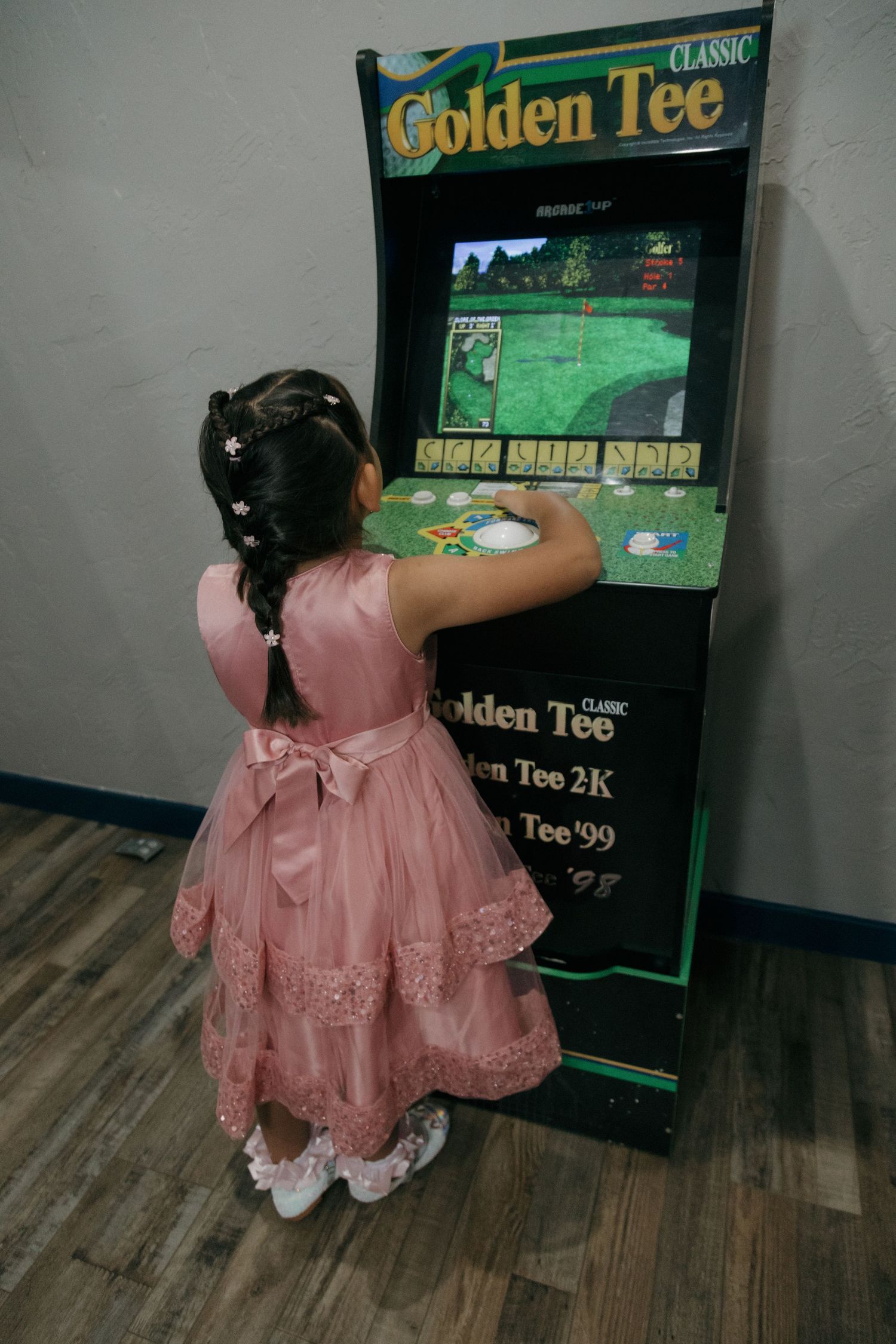 Young girl in pink dress playing Golden Tee Classic arcade game at a wedding reception venue.
