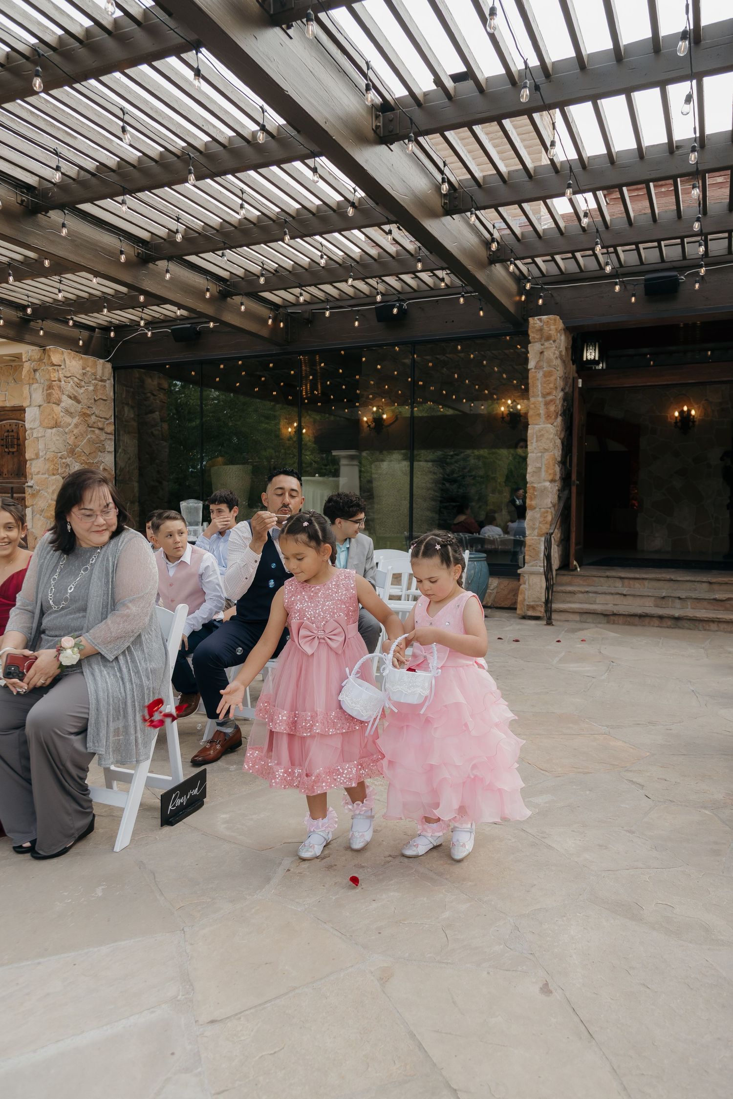 Two flower girls in pink dresses scatter petals at outdoor wedding ceremony under pergola.