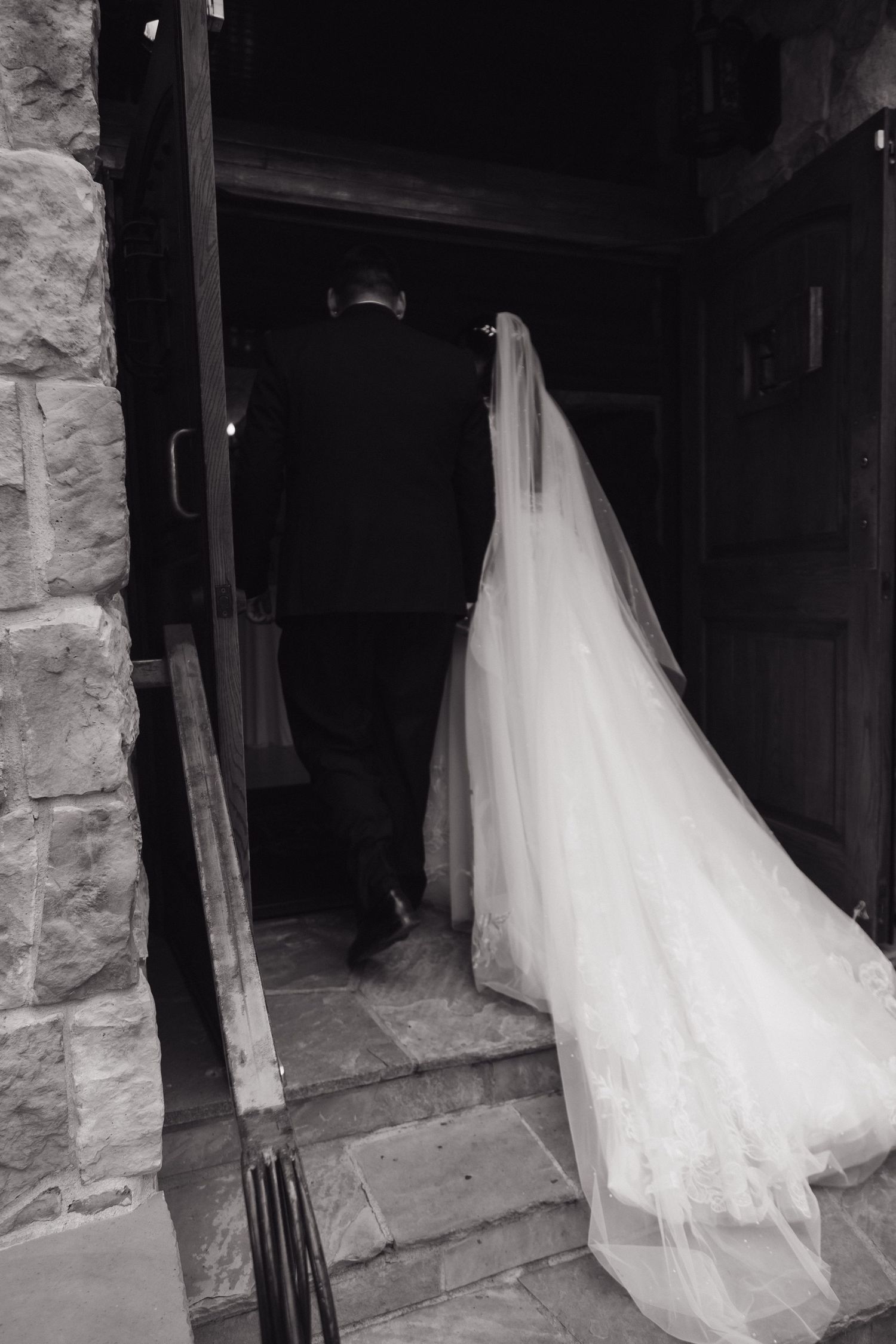 Black and white photo of a bride and groom entering a stone building, her long veil trailing behind.