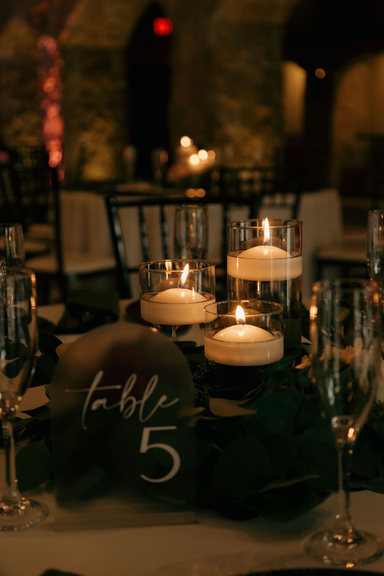 Romantic wedding reception table setting with floating candles and a dark green Table 5 sign in candlelight.