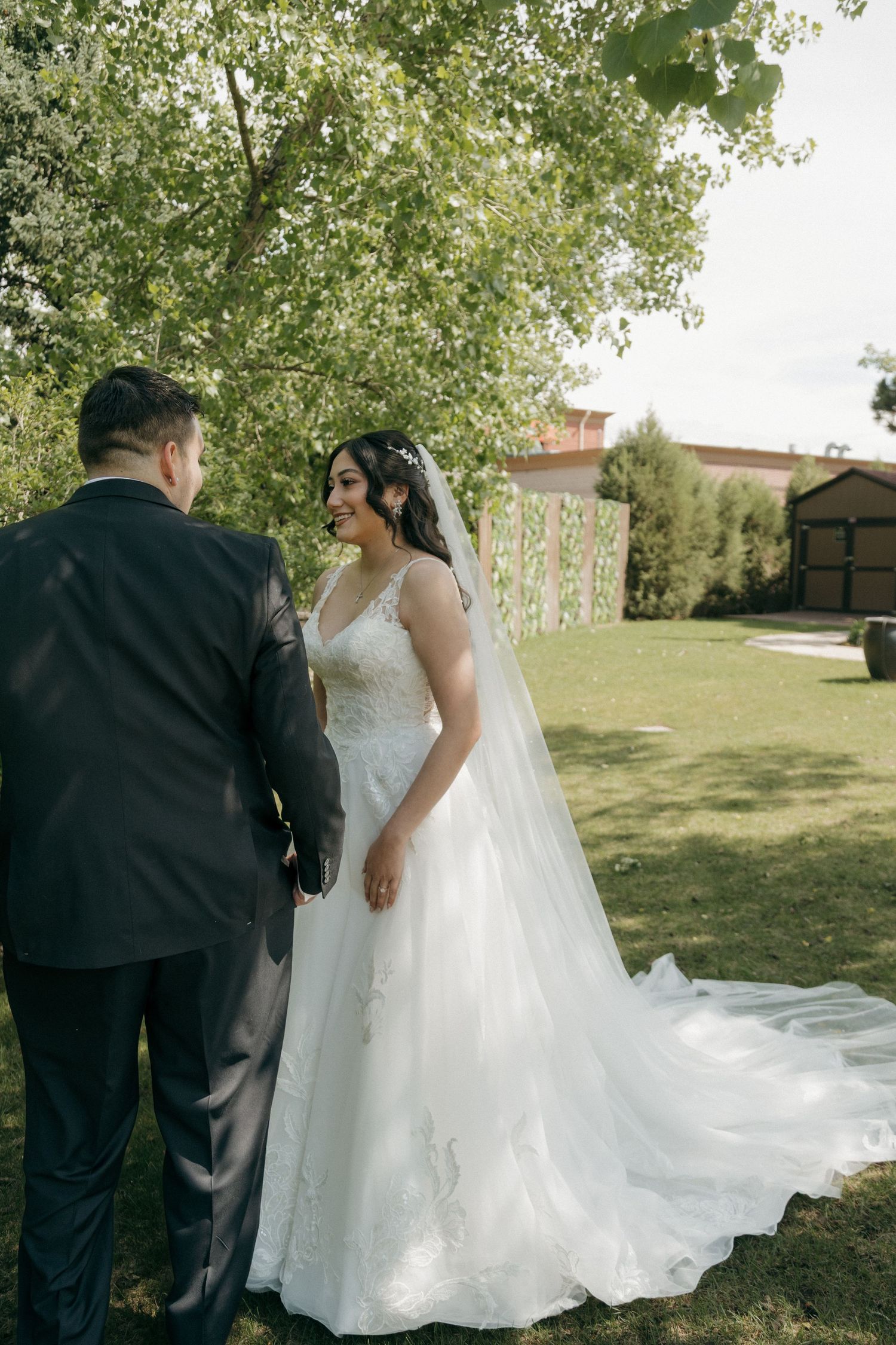 Bride in a lace ballgown with long veil smiling at groom during outdoor wedding portraits under green trees.