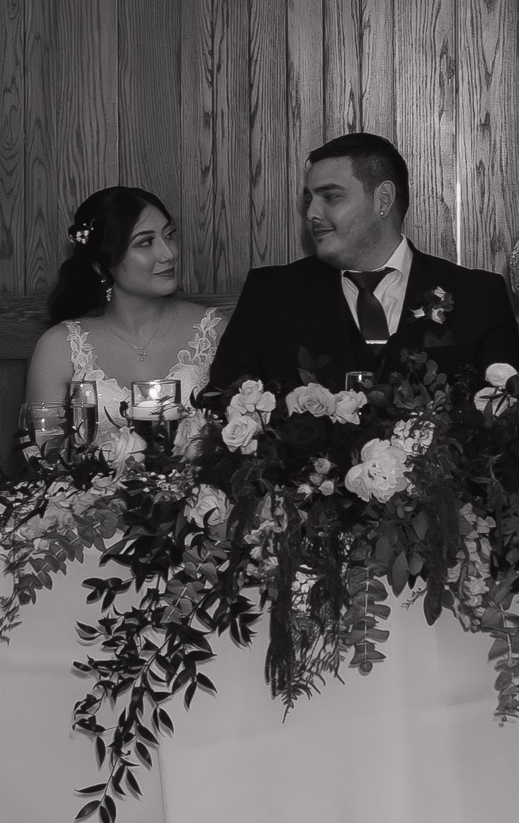 Black and white portrait of bride and groom at sweetheart table surrounded by elegant floral arrangements.