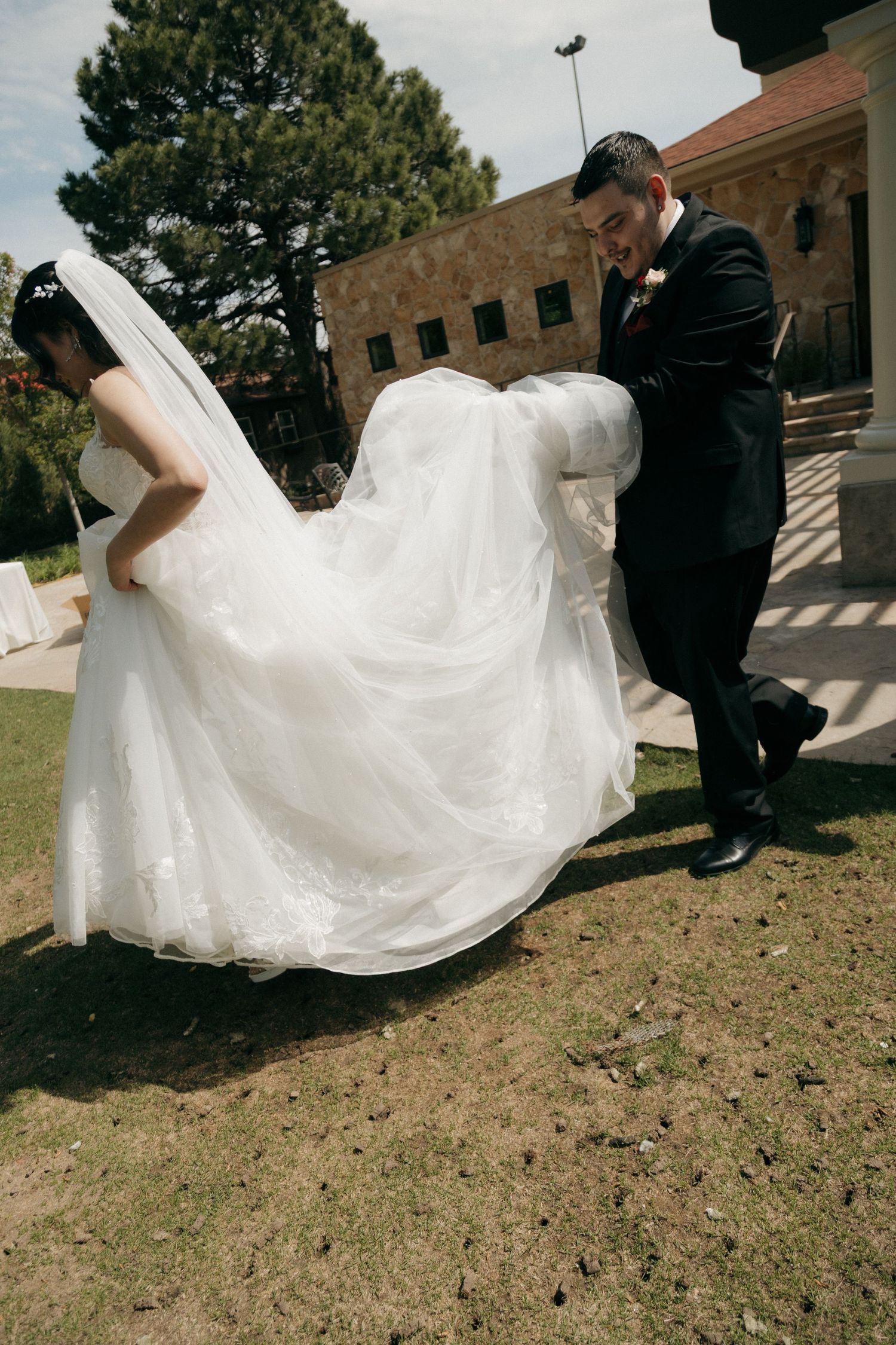 Groom helping bride carry her white lace wedding dress train across green lawn outside a stone venue.
