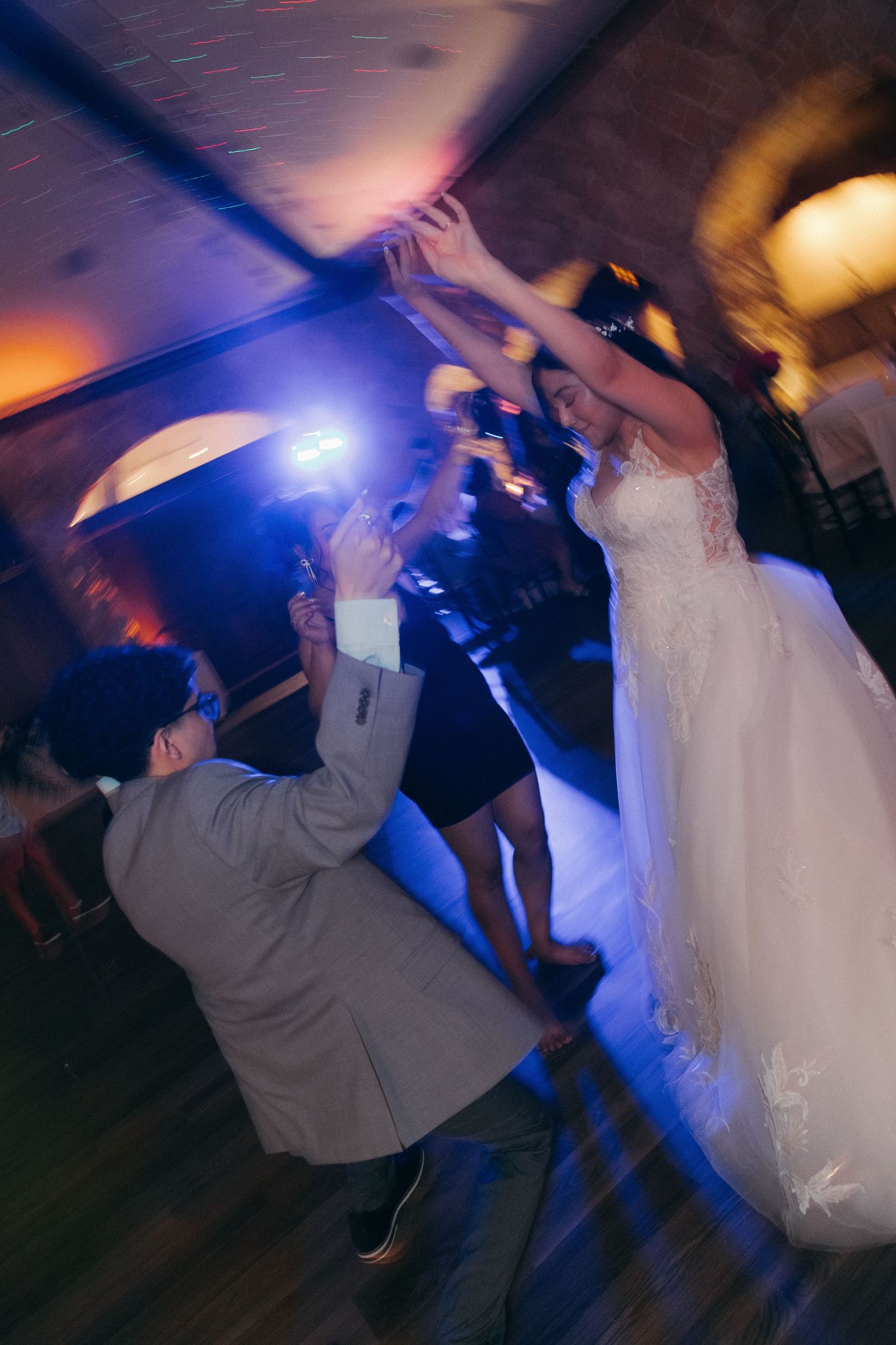 Bride in lace gown dances joyfully with guest under blue lights at vibrant wedding reception.