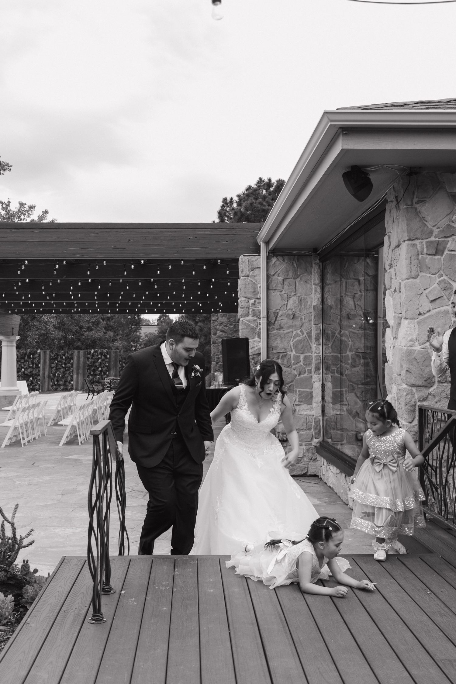 Black and white photo of bride and groom walking on deck with flower girls playing nearby at outdoor wedding venue.