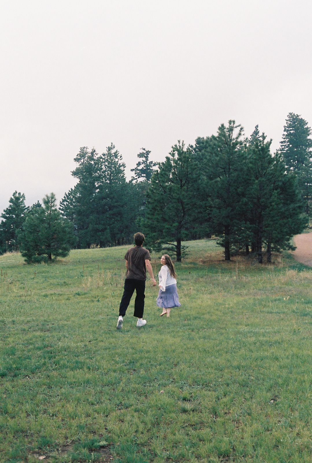 Couple holding hands while walking through a lush green meadow surrounded by tall evergreen trees.