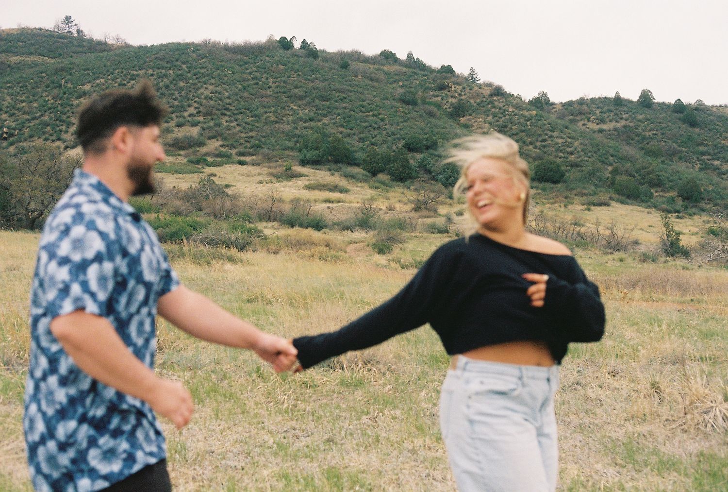 A couple holds hands and dances in a green meadow with rolling hills, the woman laughing as wind blows her blonde hair.