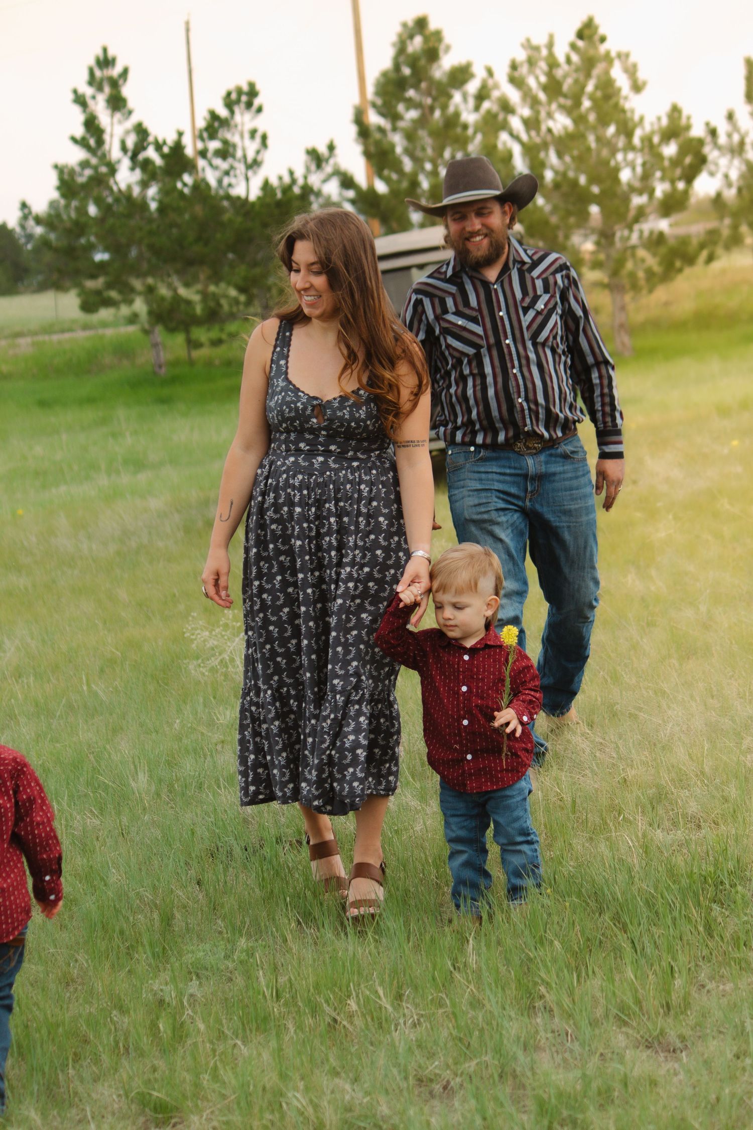 Family takes a casual stroll together through a green field with pine trees in background.