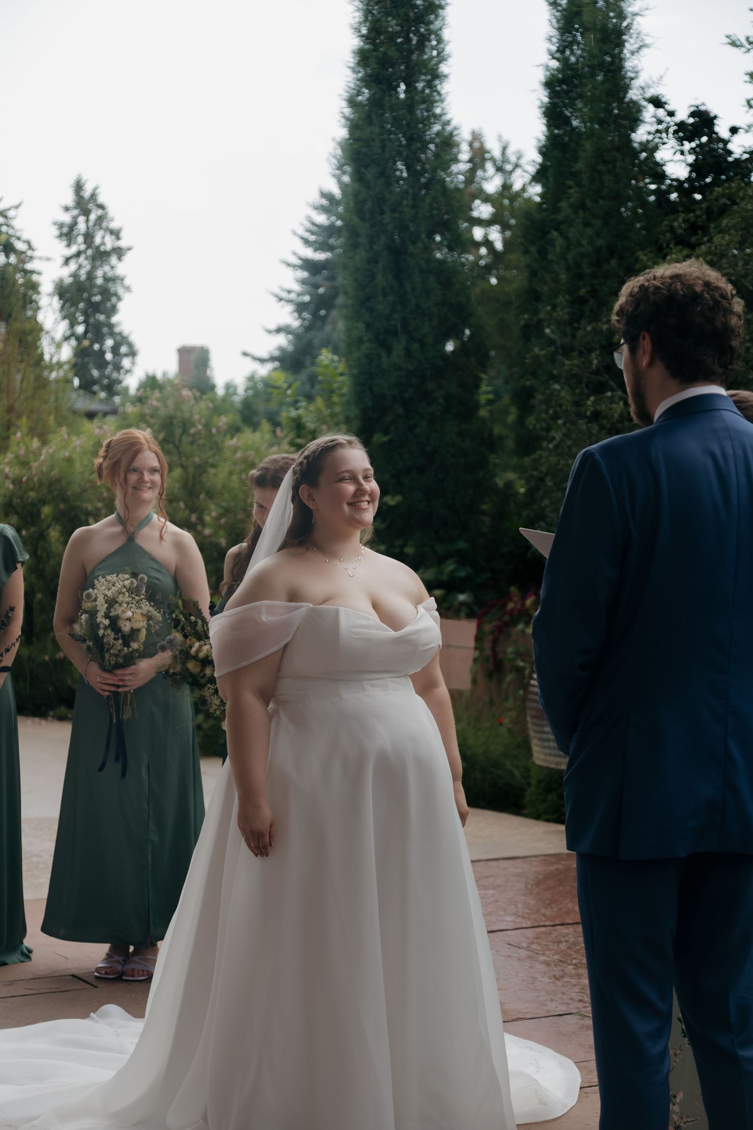 Radiant bride in off-shoulder white gown smiling at groom during outdoor wedding ceremony with bridesmaids in green.