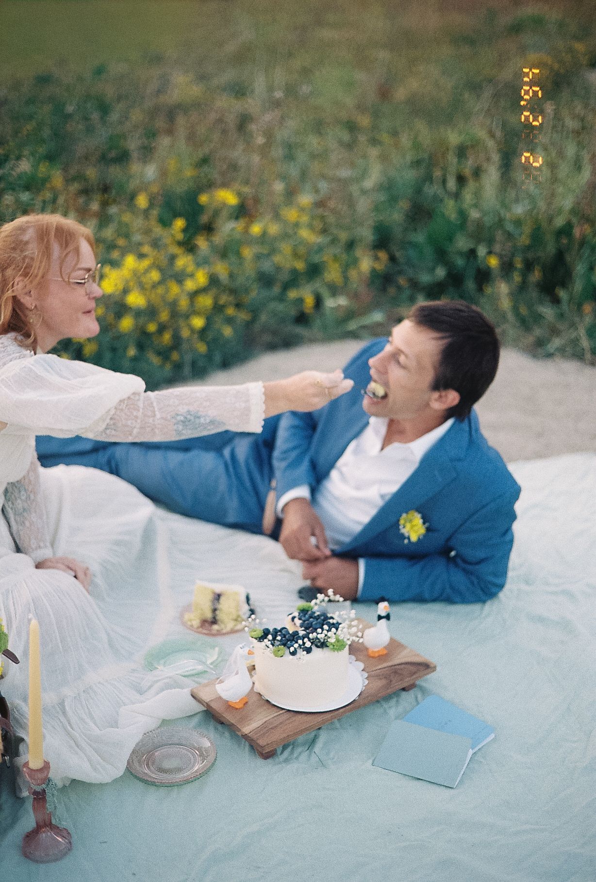 Bride feeds groom wedding cake during an outdoor picnic celebration with wildflowers in the background.