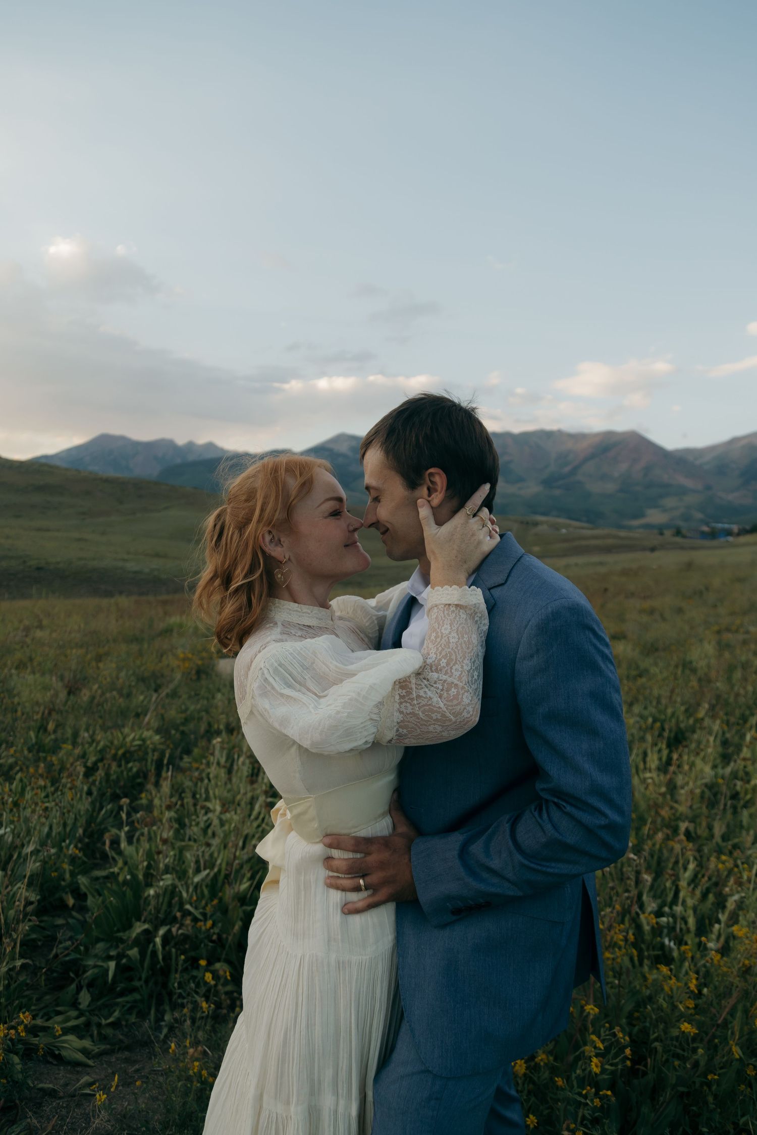 Bride and groom share romantic almost-kiss in lush green mountain meadow at dusk during outdoor wedding.